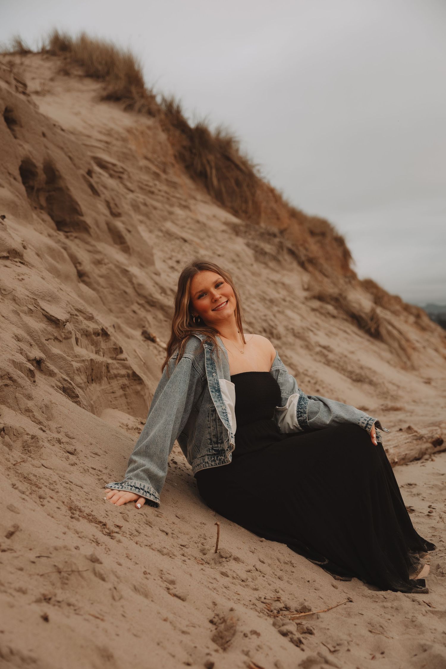 A person in a black dress and denim jacket reclines on sandy dunes against an overcast sky.