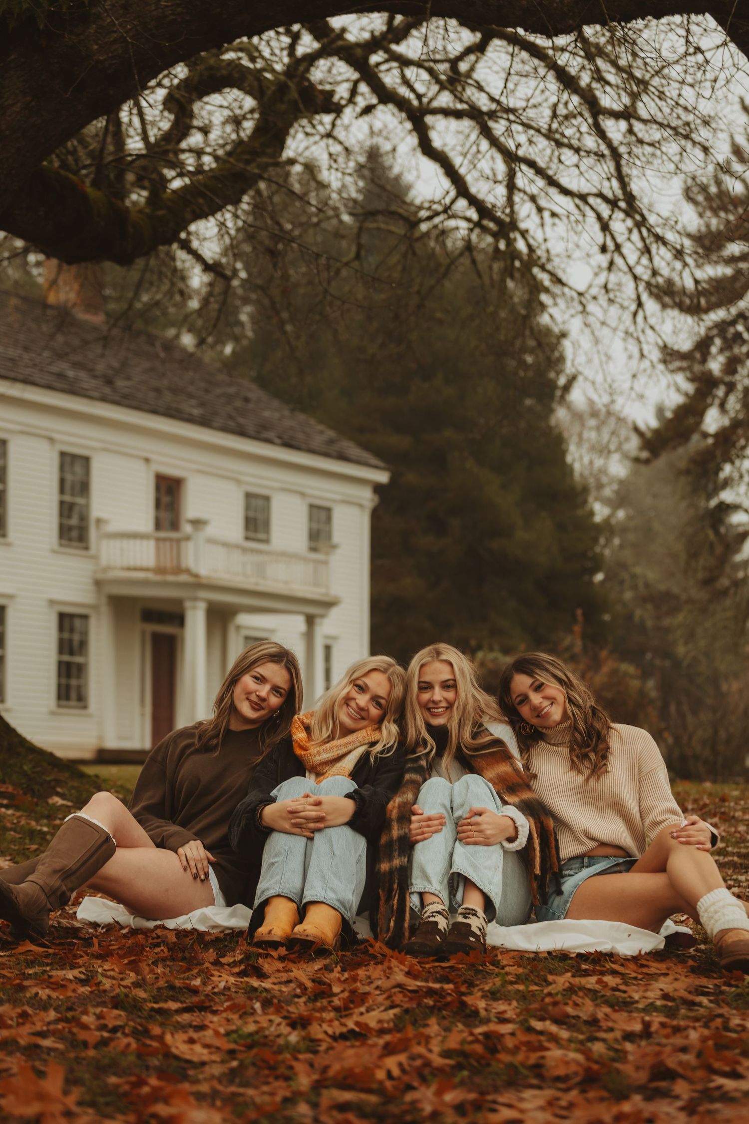 A group of friends lying together in autumn leaves outside a white colonial style house on a fall day.