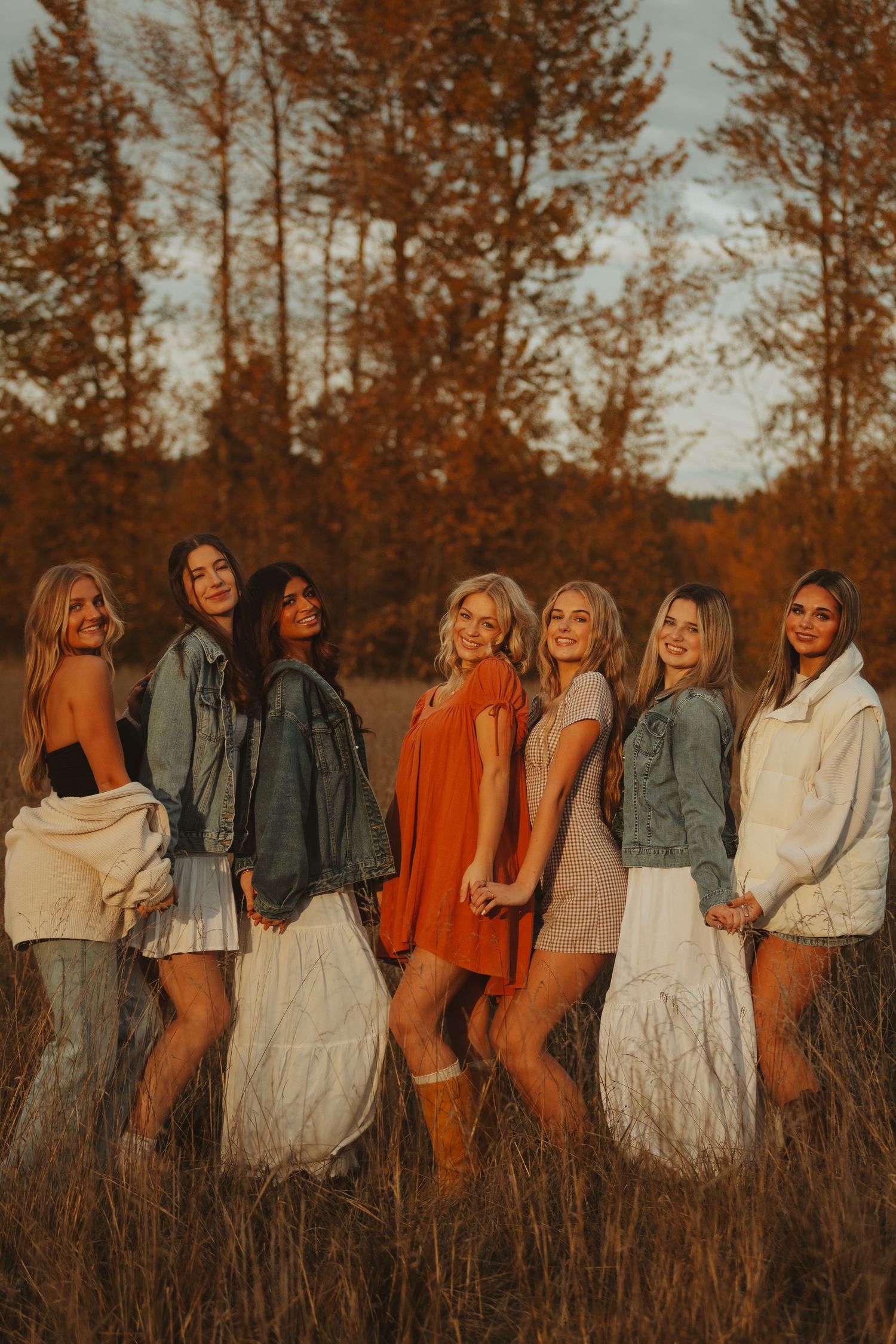 A group poses together in white dresses and denim jackets in a fall field at golden hour.