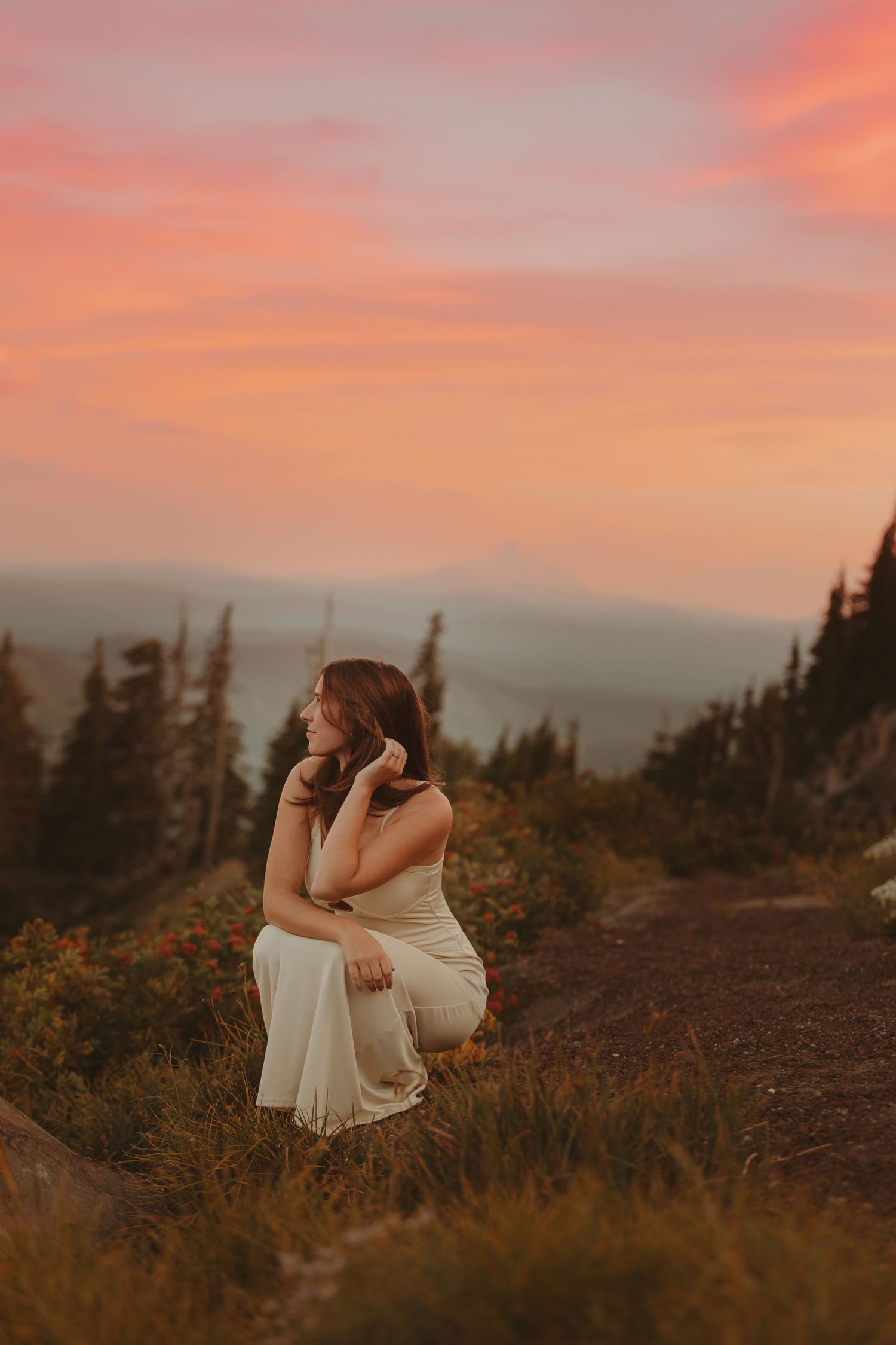 Woman in white dress crouching on mountain trail at sunset with pink sky and forest valley backdrop.
