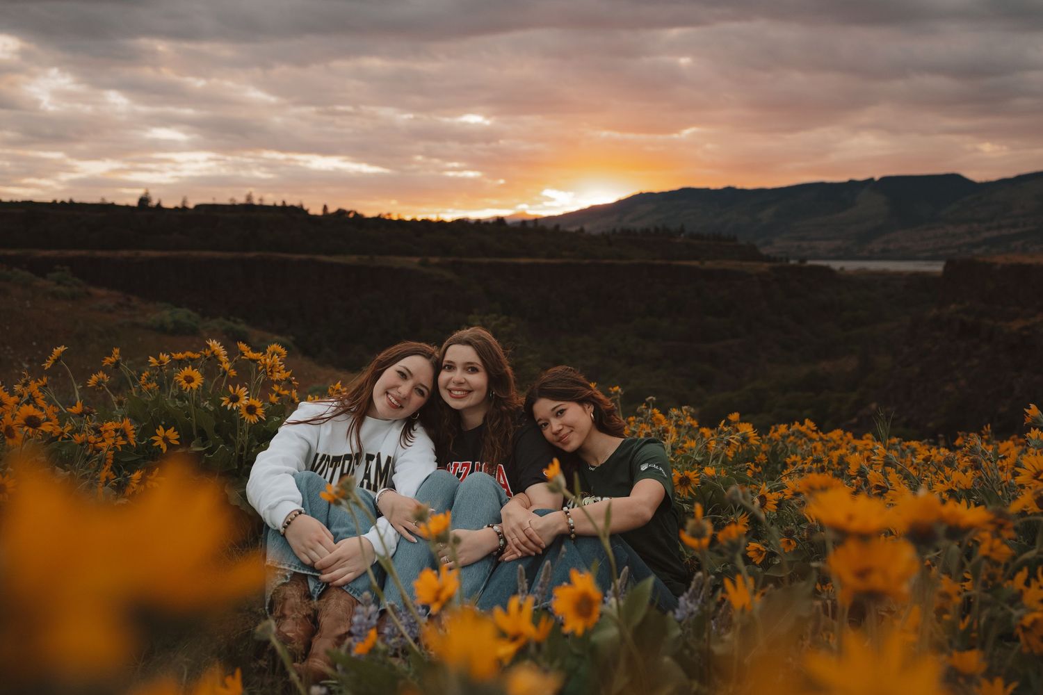 Three young women smile together in a field of yellow wildflowers at sunset with mountains in the background.