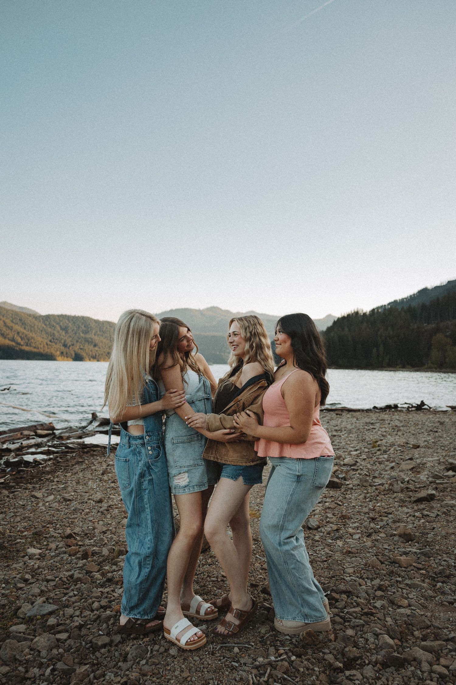 Four young women laughing and embracing on a rocky lakeside with mountains in the background at dusk.