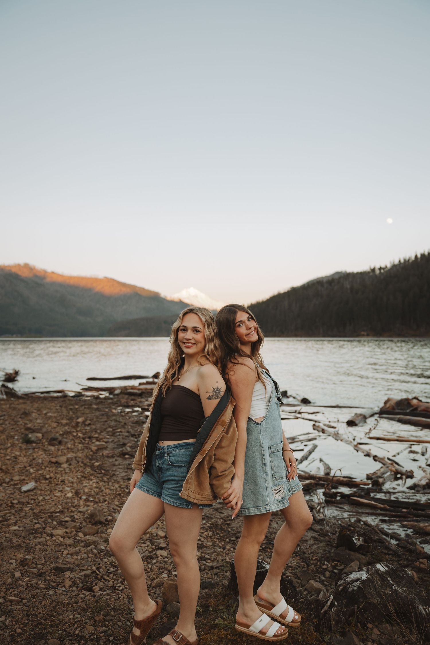 Two smiling young women posing back-to-back on a rocky lakeshore at sunset.