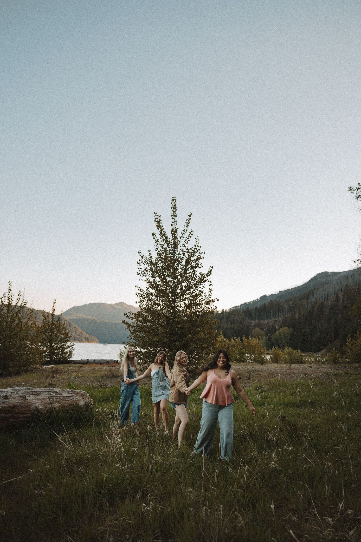 Four young women holding hands walking through a meadow near a mountain lake at dusk.