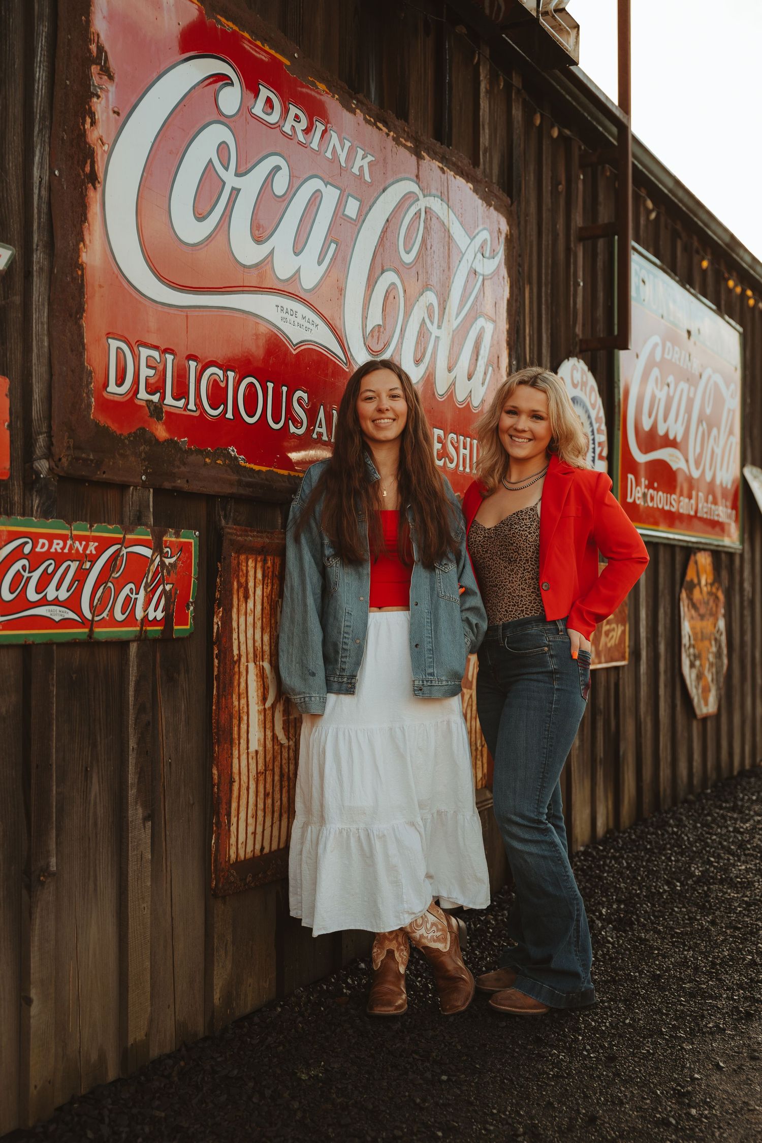 Two women in western attire smiling beside vintage Coca-Cola signs on a rustic wooden wall.