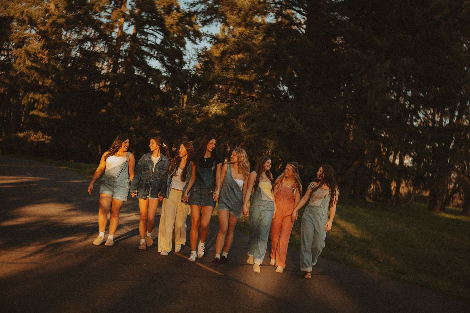 A group of young women walking together on a sunlit road surrounded by lush green trees during golden hour.