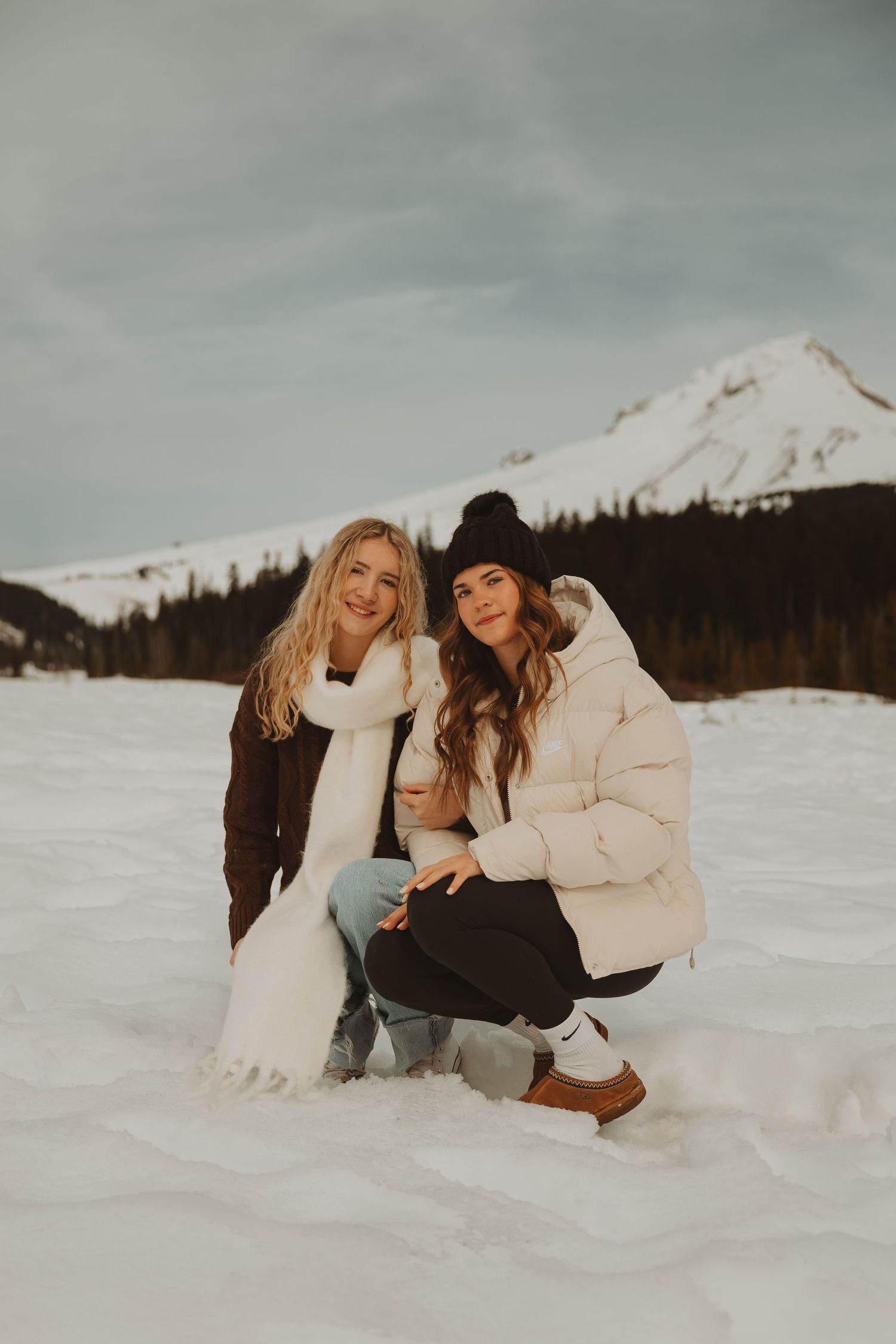 Two women pose in snow with mountain backdrop wearing winter coats and scarves.