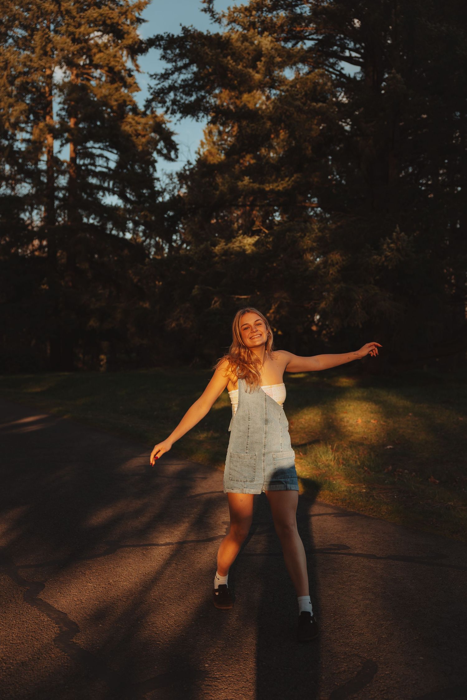 Joyful young woman in denim overalls dancing on a road at golden hour surrounded by tall trees.