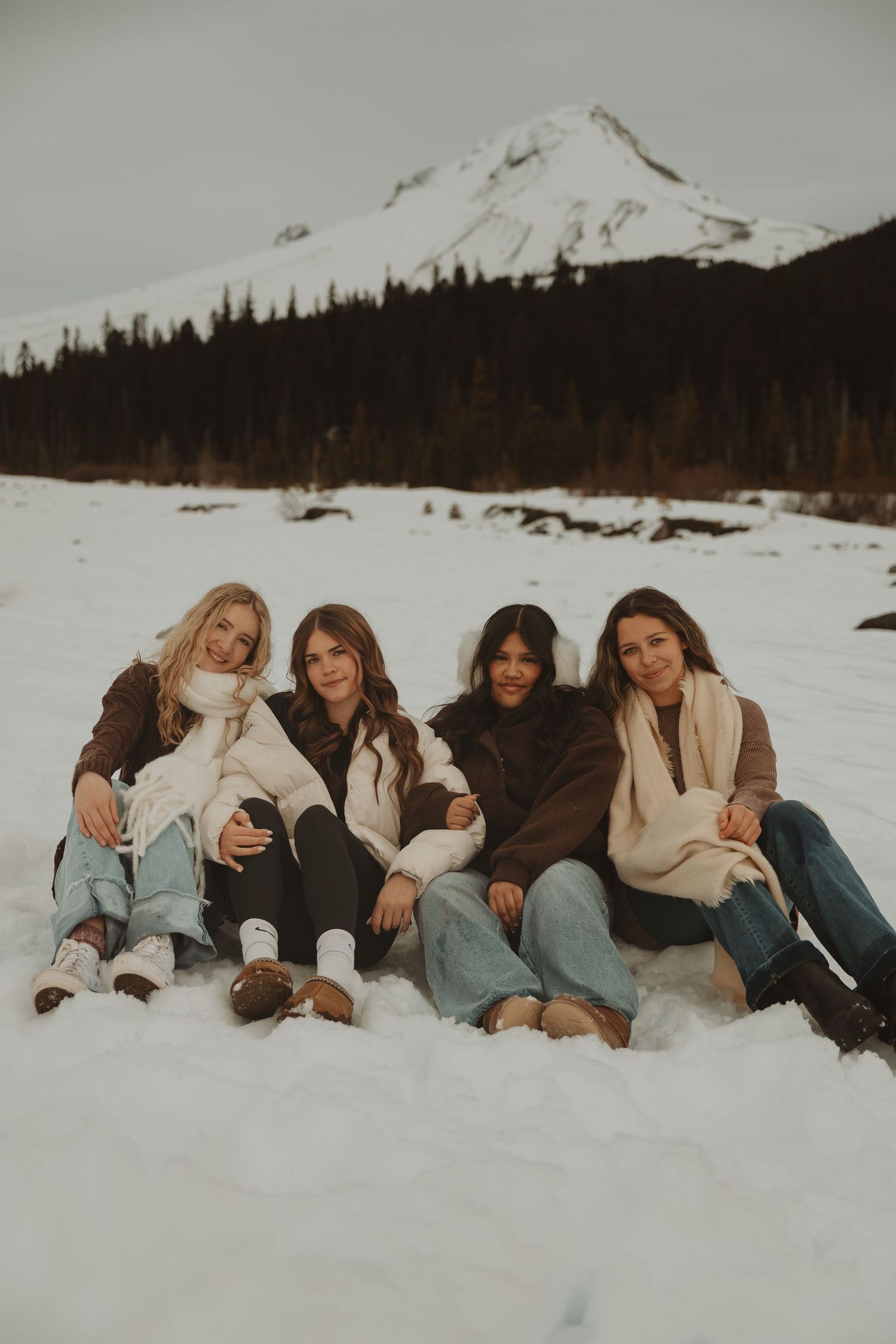 Four women sitting together in snow smiling with mountain peak backdrop on overcast day.