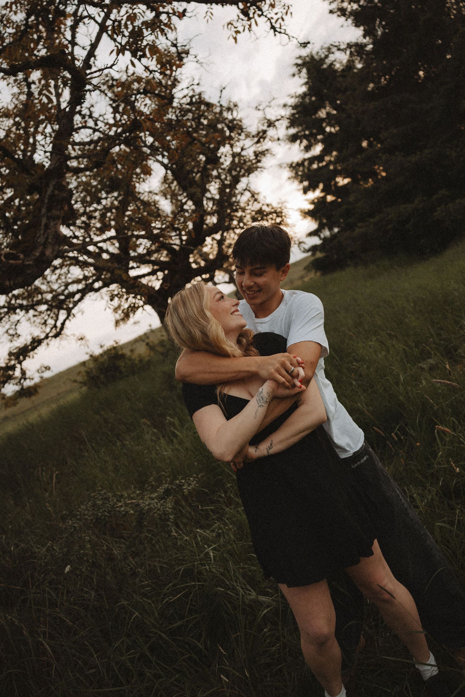 Happy couple embracing and laughing together in a green field surrounded by trees at dusk.
