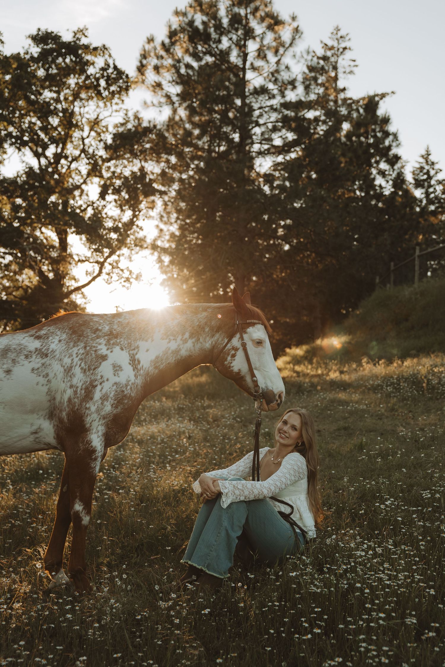 Smiling young woman sits beside a brown and white horse in a golden sunset meadow with wildflowers.
