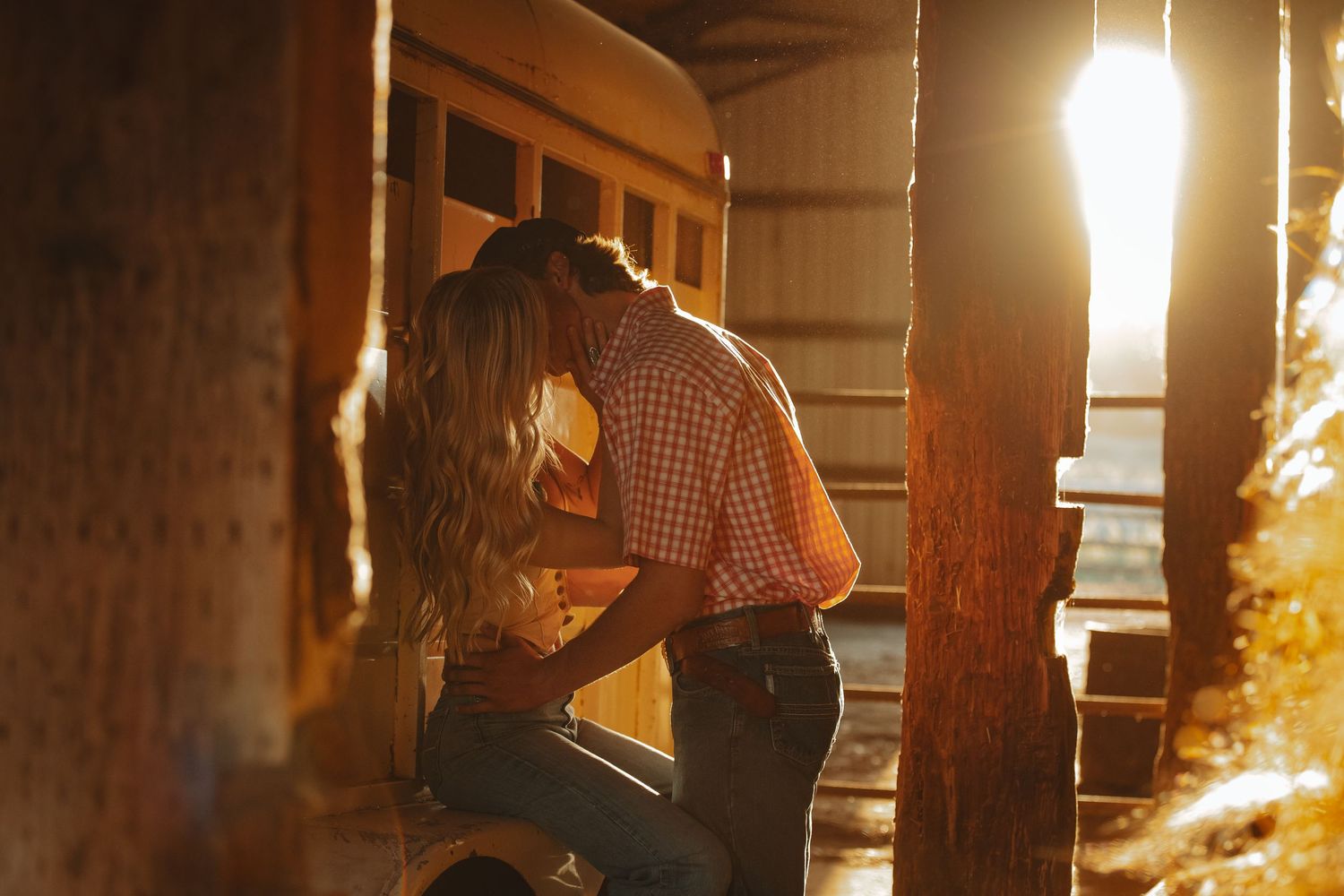 Couple shares intimate moment leaning against barn wall in warm sunset glow.