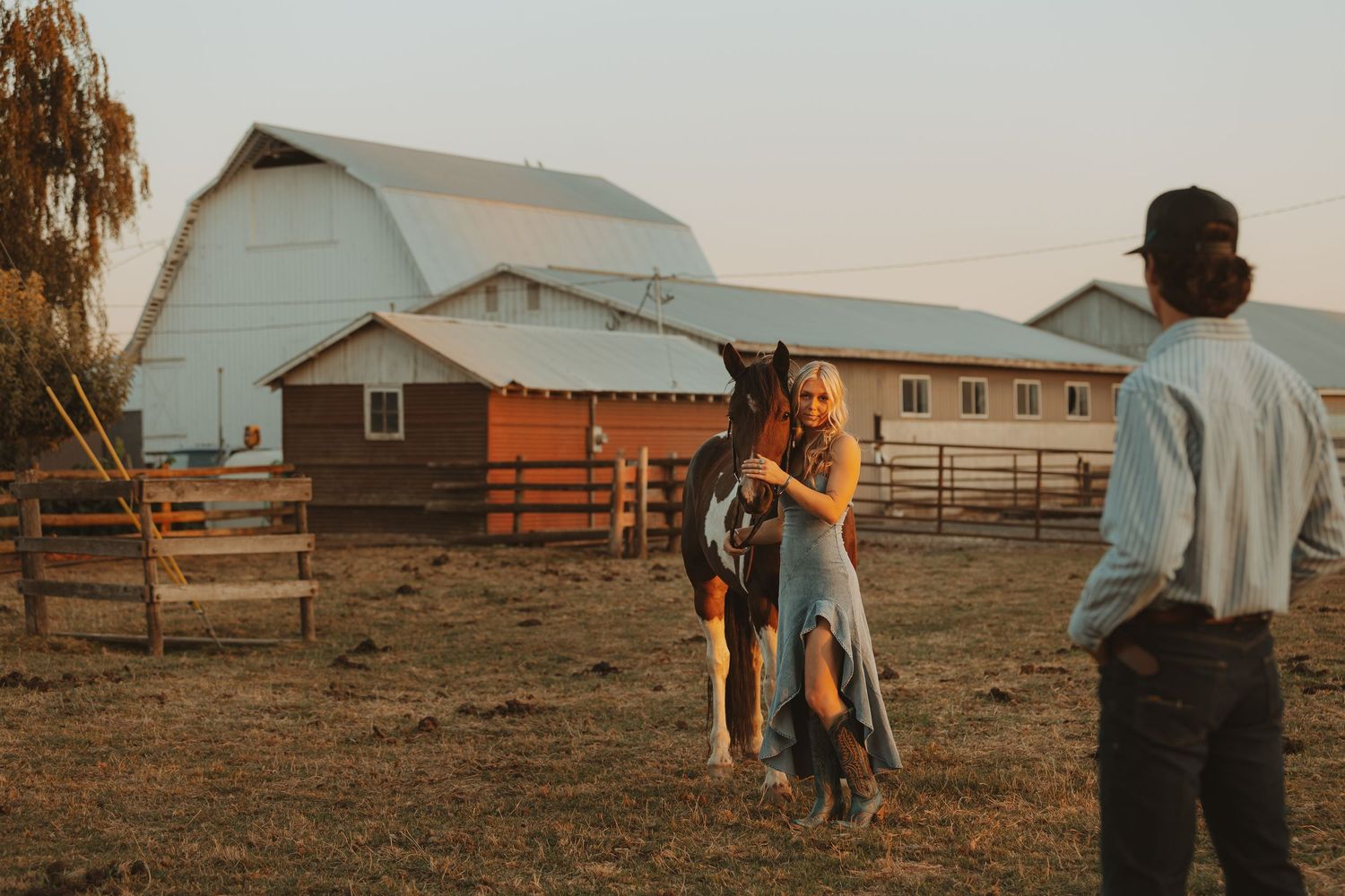 Couple poses with a horse at golden hour on a rustic farm with a white barn in the background.
