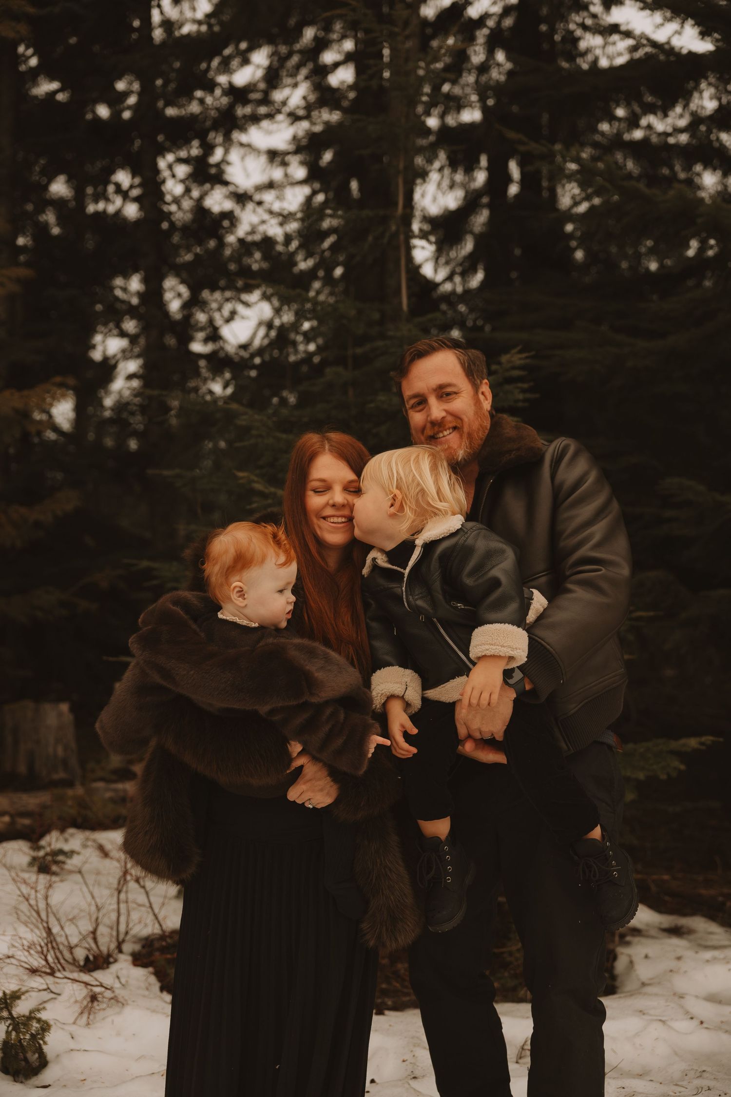 Family sharing tender moments together in a snowy winter setting with evergreen trees in the background.