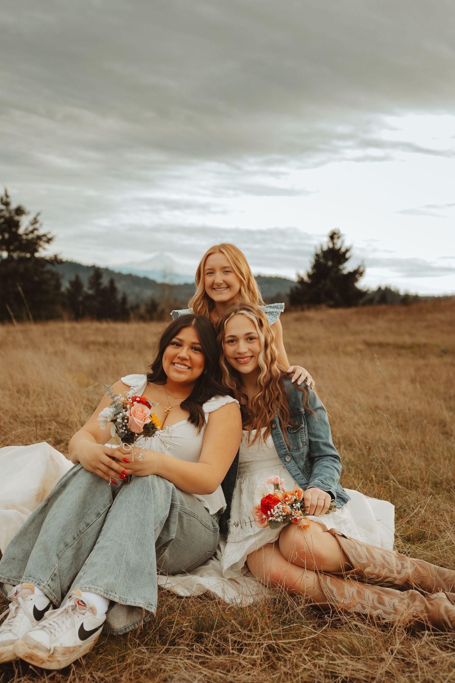 Friends lounging together in a golden field wearing denim and white dresses with floral bouquets.