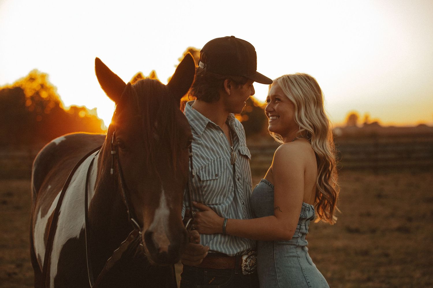 Smiling couple standing close together with a paint horse at golden hour on a farm.