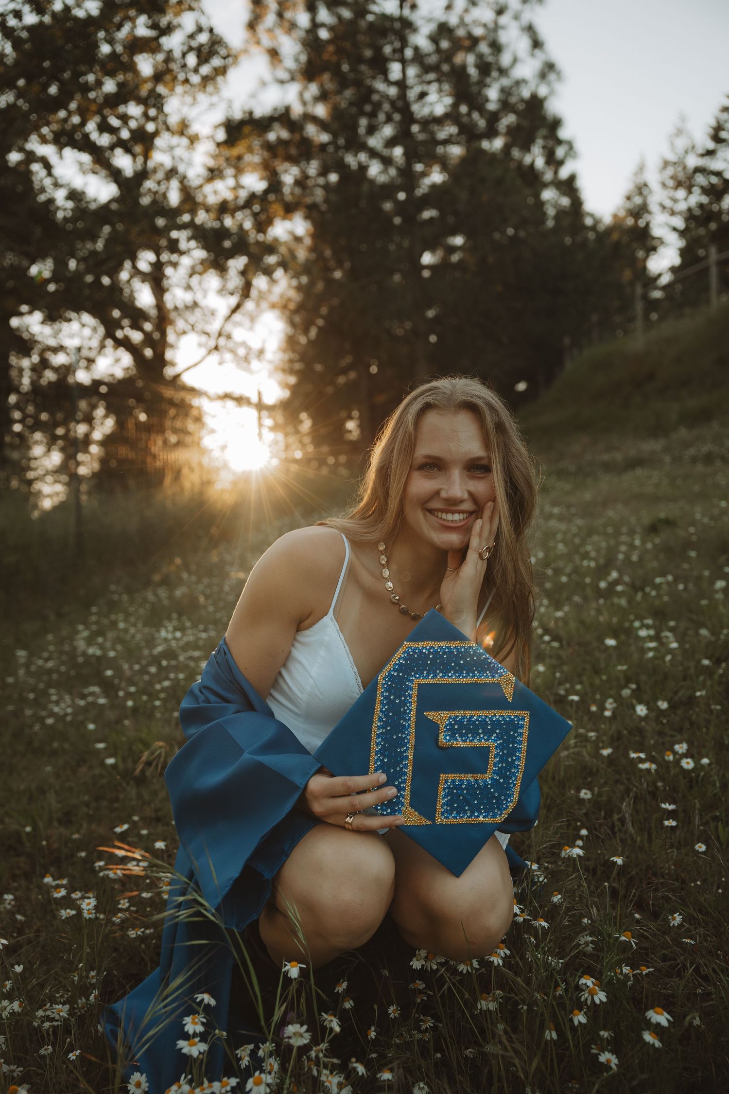 Happy young woman in white dress displays decorated blue graduation cap in flower field at dusk.