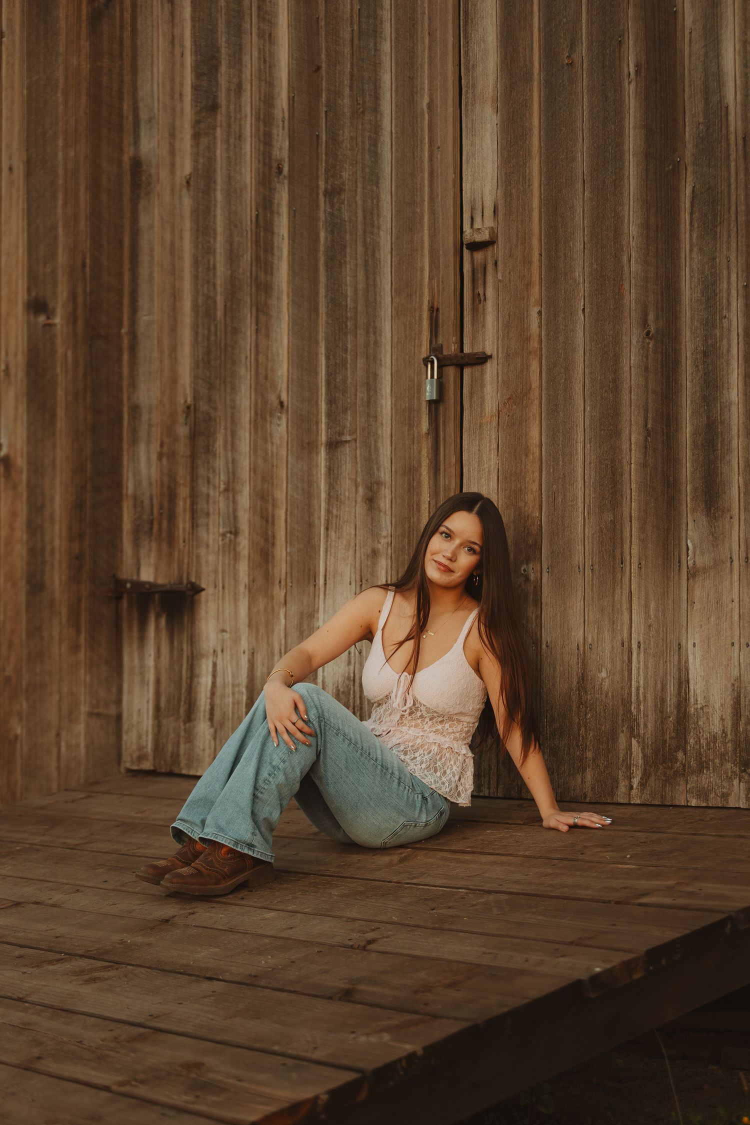 Young woman in flare jeans and white top sitting against rustic wooden barn wall on a wooden deck.
