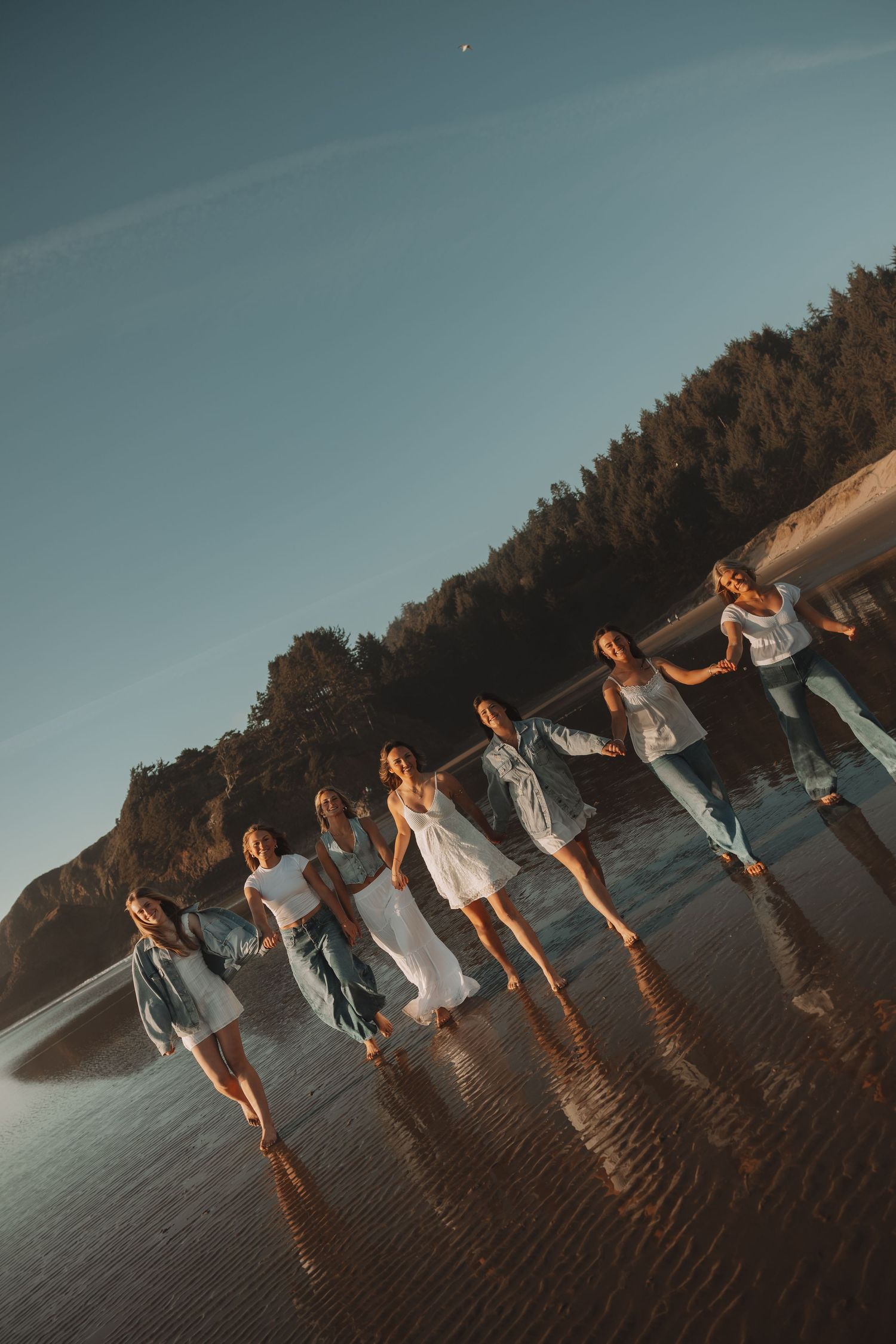 Group of friends holding hands walking along a reflective beach shoreline at golden hour sunset.