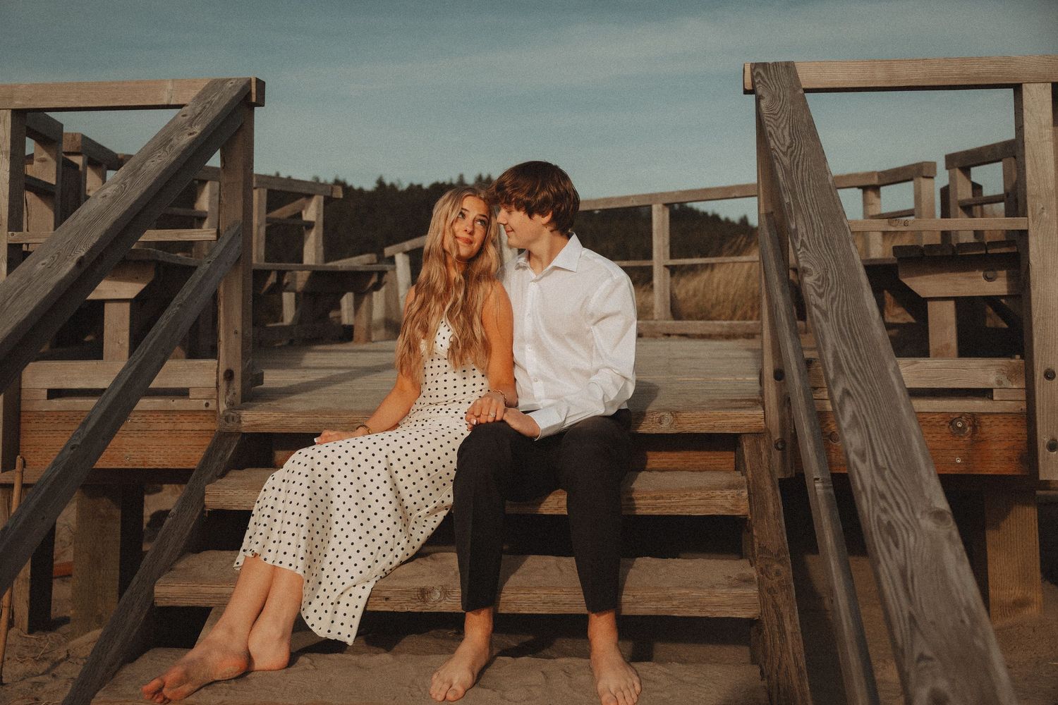 Couple sitting together on wooden beach boardwalk stairs, woman in floral dress, man in white shirt, sharing a romantic moment.