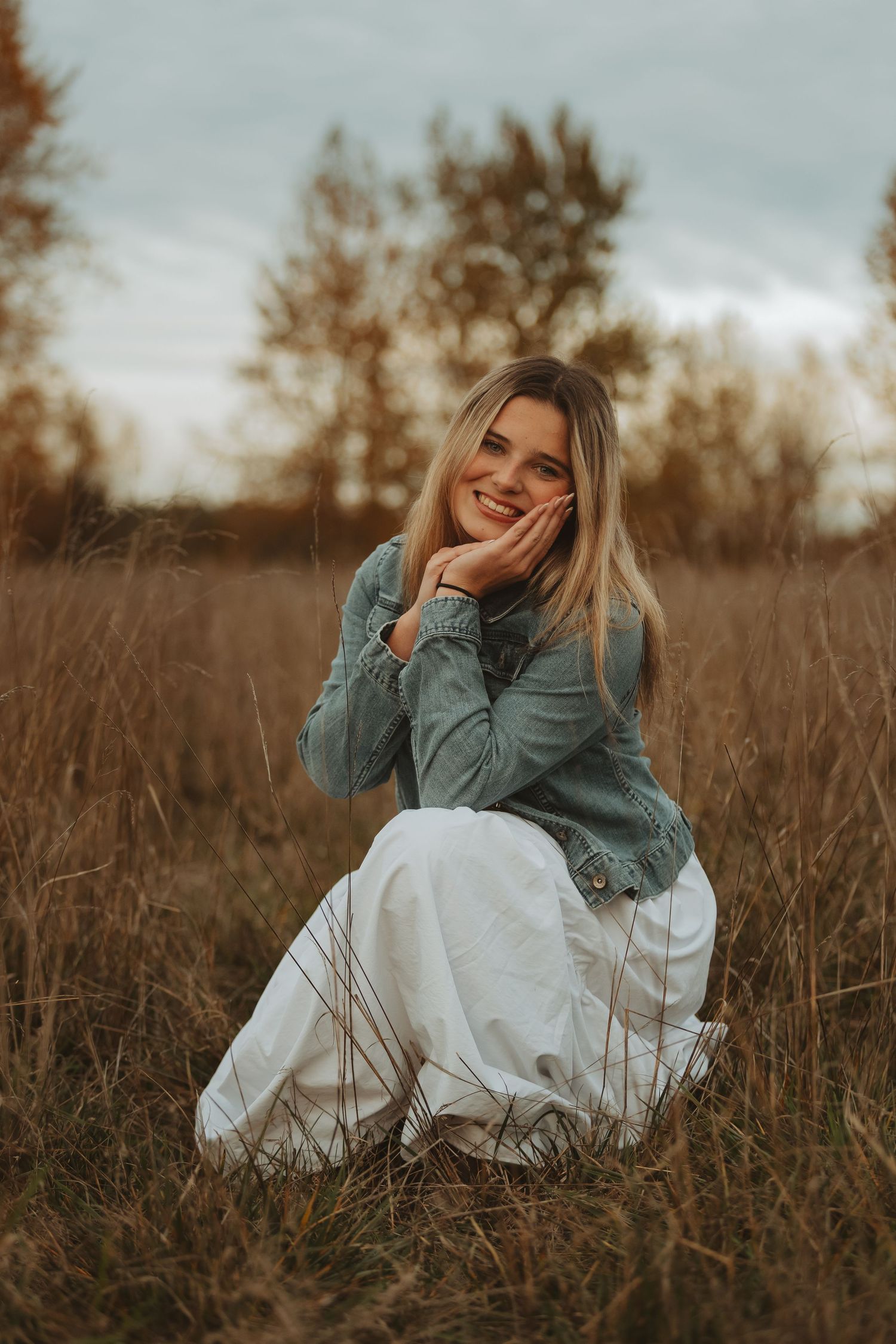 Figure in denim jacket and white dress reclines in tall grass during sunset photoshoot.