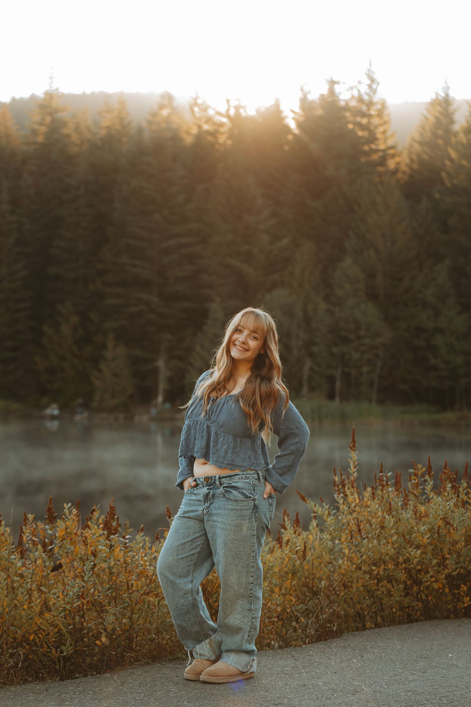 Person in denim overalls dances joyfully on a path at sunset with evergreen trees and golden wildflowers in background.