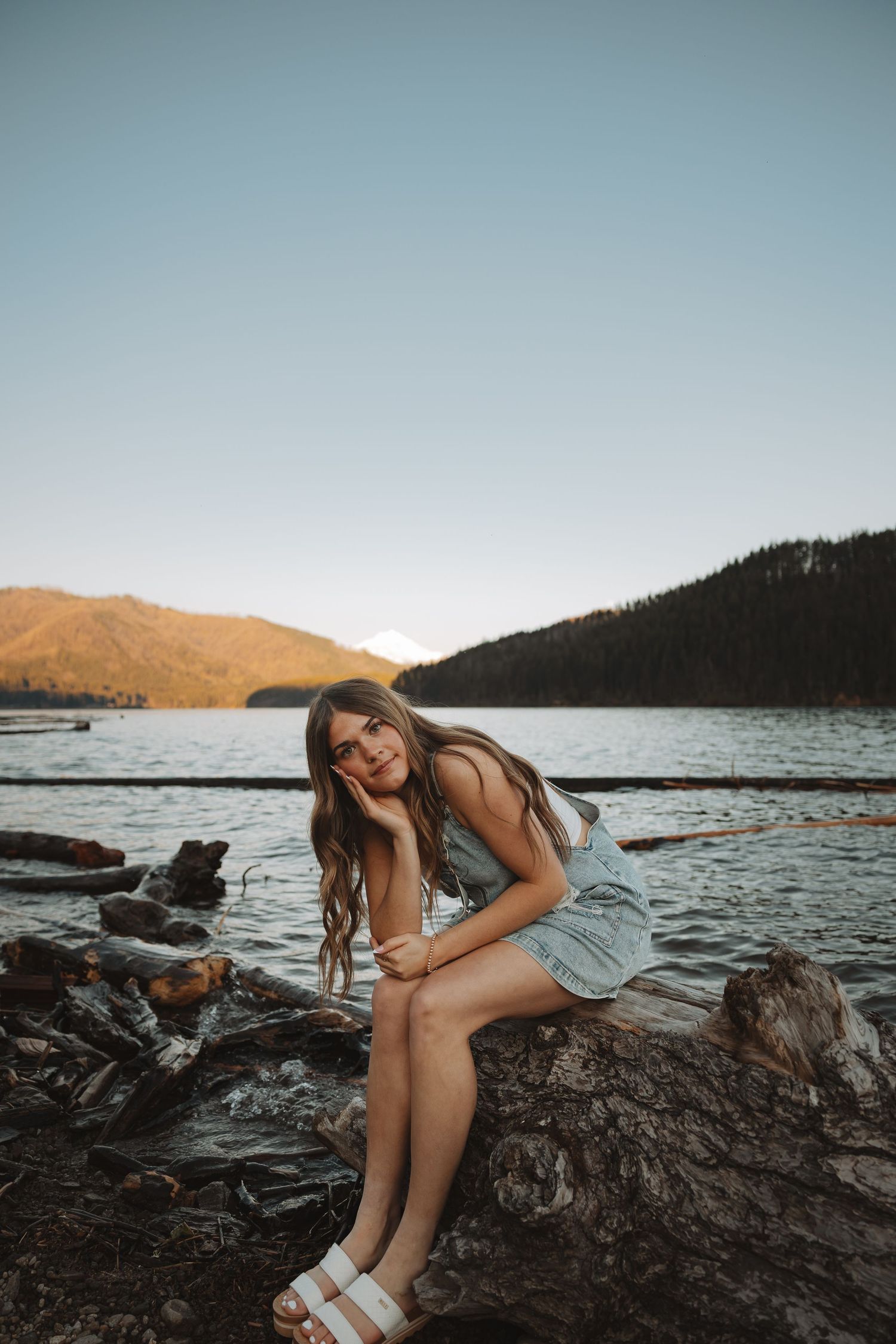 Young woman sitting on rocks by a serene mountain lake at sunset, wearing a light blue romper and white sandals.