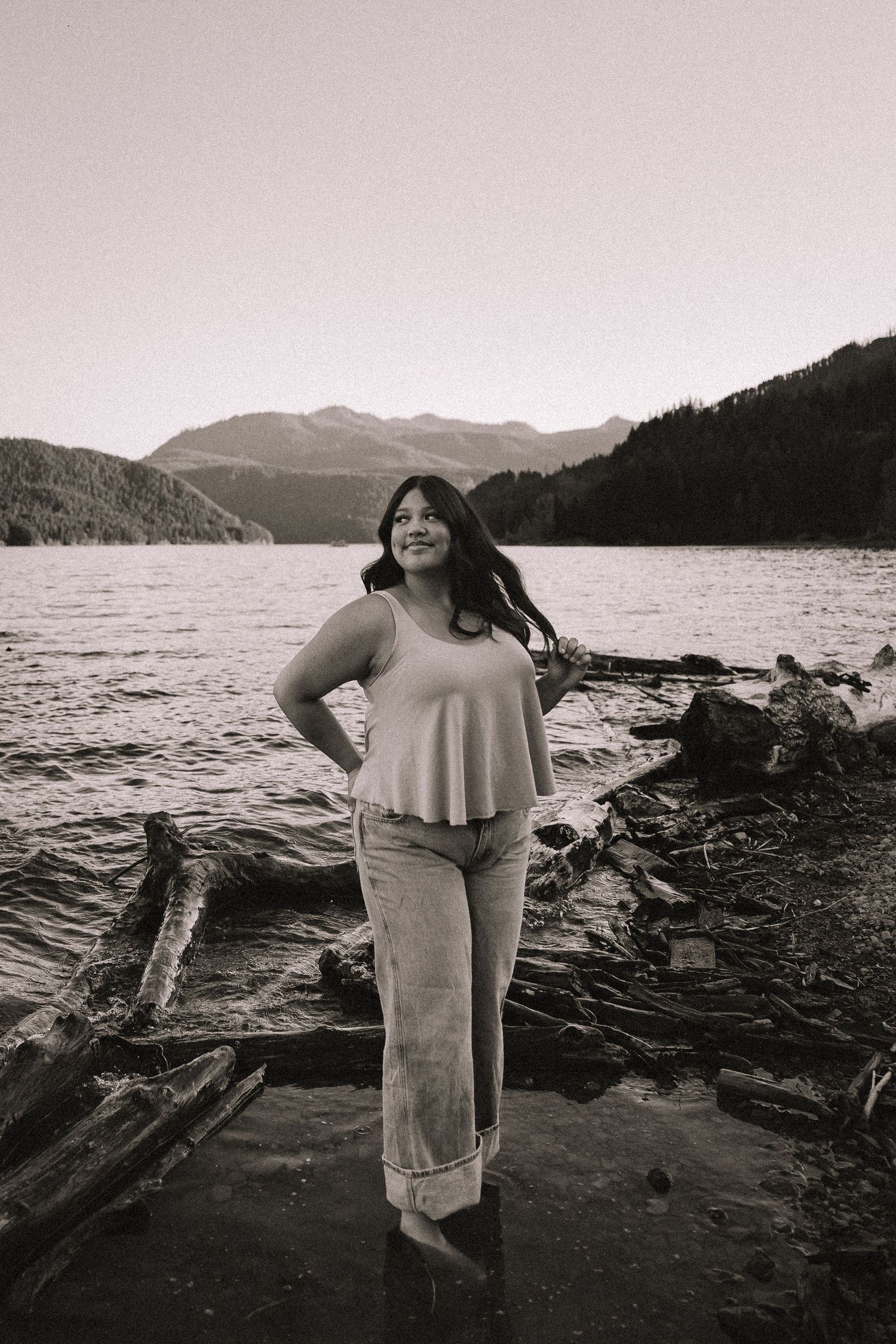 Black and white portrait of a smiling woman standing on driftwood by a mountain lake.
