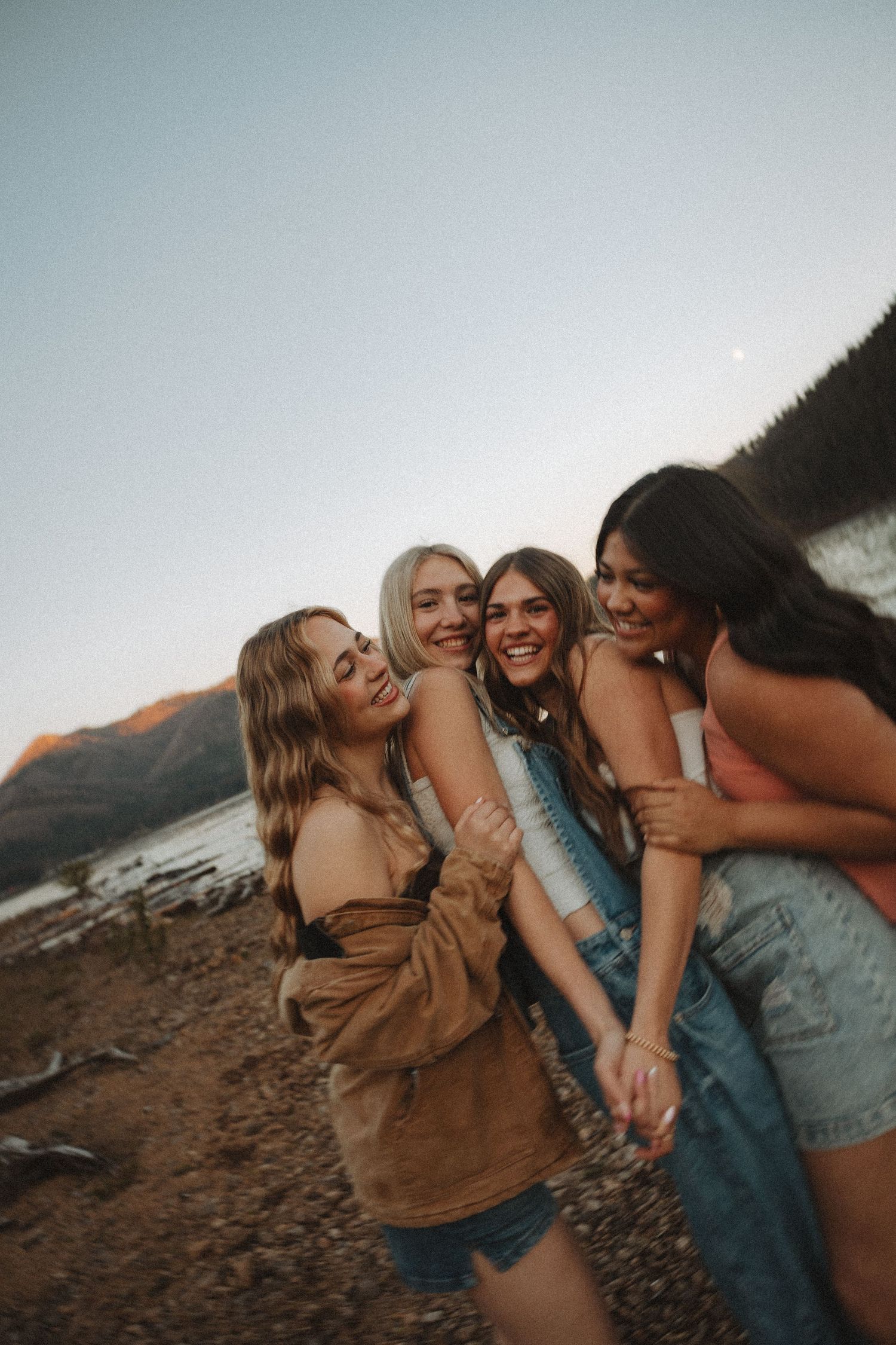 Four young women laughing and embracing on rocky lakeshore with mountains and crescent moon at dusk.
