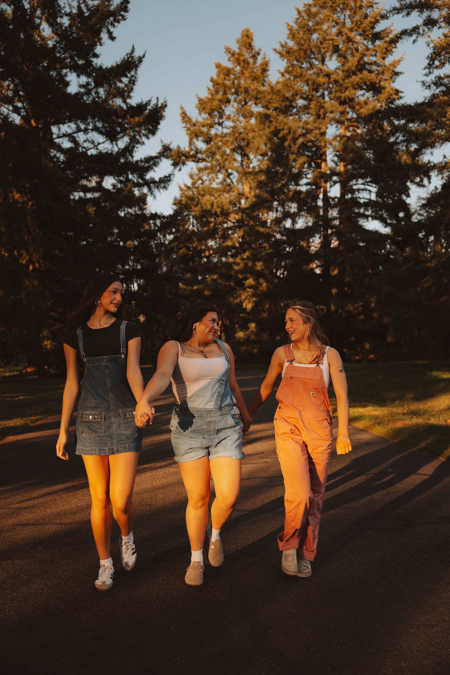 Three young women in overalls walking hand-in-hand on a road surrounded by tall pine trees at sunset.