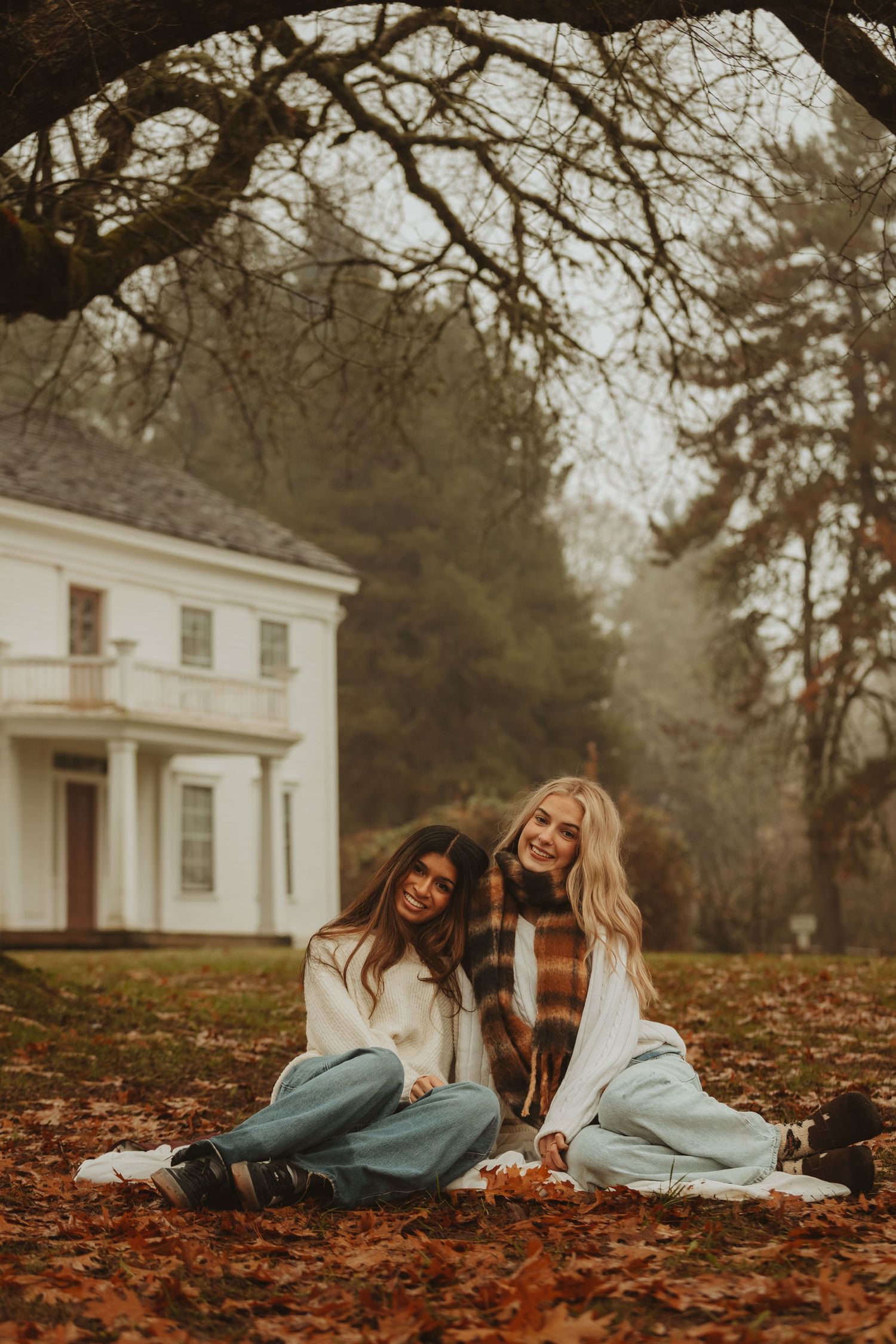 Two friends in white sweaters sit laughing together on autumn leaves in front of a white house on a misty day.