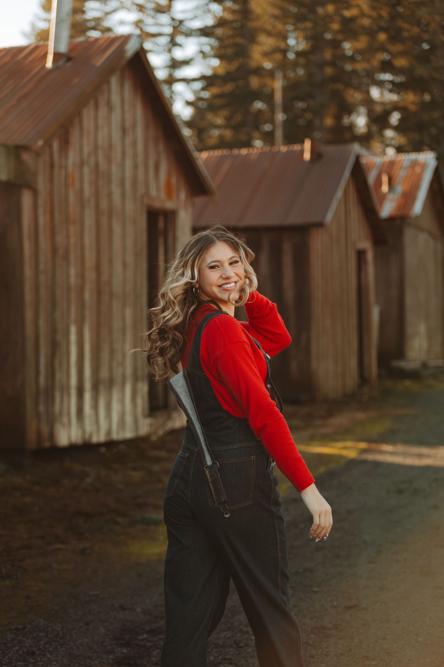 A person in a red sweater and overalls poses playfully in front of rustic wooden buildings at sunset.