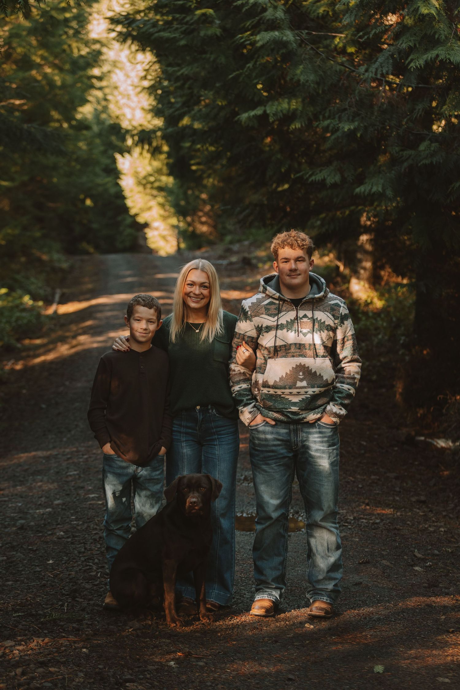 A family and their Rottweiler dog walk together on a wooded path during golden hour.