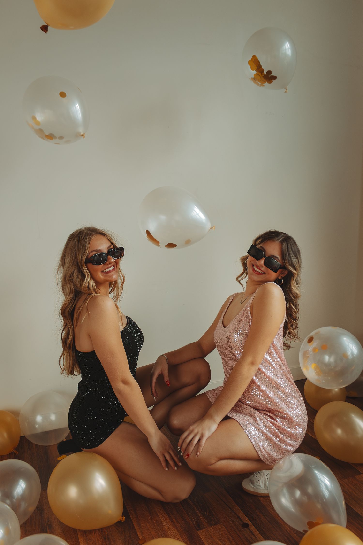 Two people in party outfits posing with gold and silver balloons on hardwood floor.