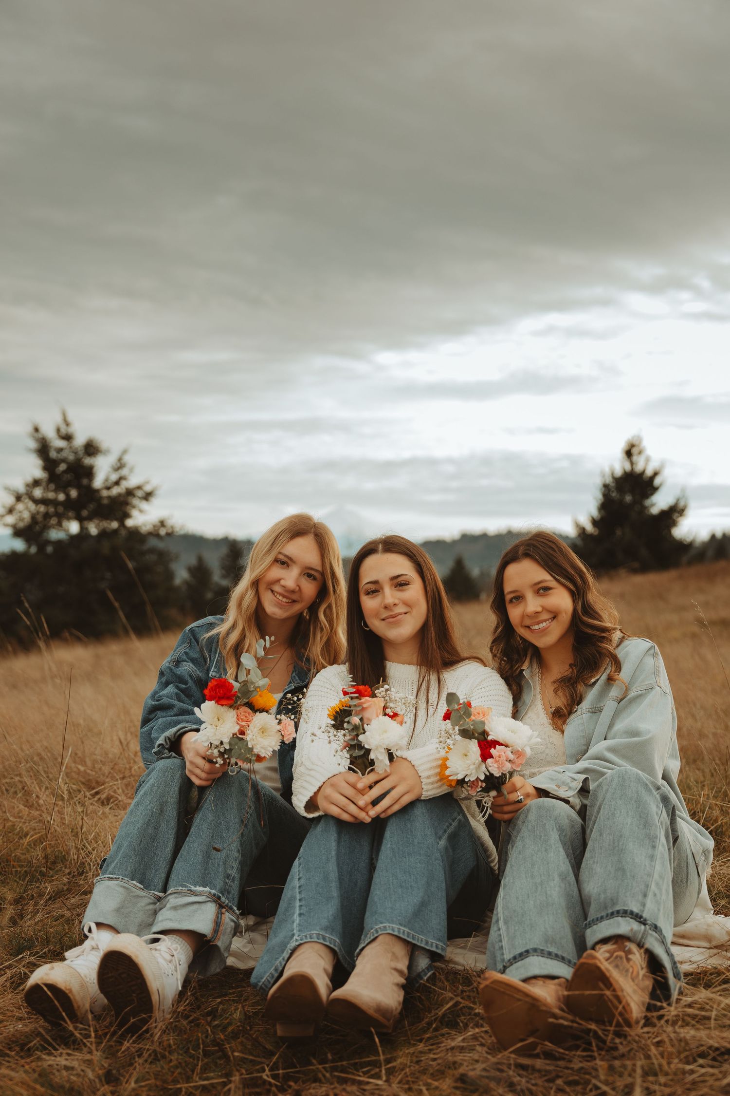 Friends in matching denim outfits lay in tall grass holding flowers under cloudy skies.