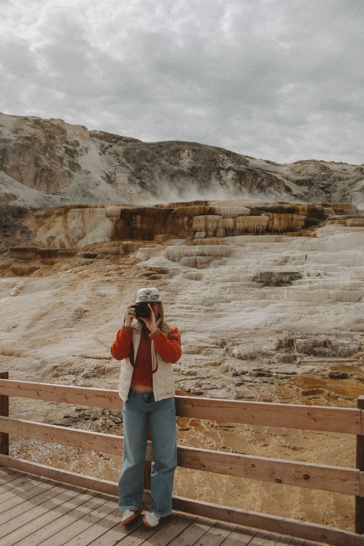 A woman photographs Mammoth Hot Springs terraces at Yellowstone, wearing jeans and a red jacket on a wooden boardwalk.