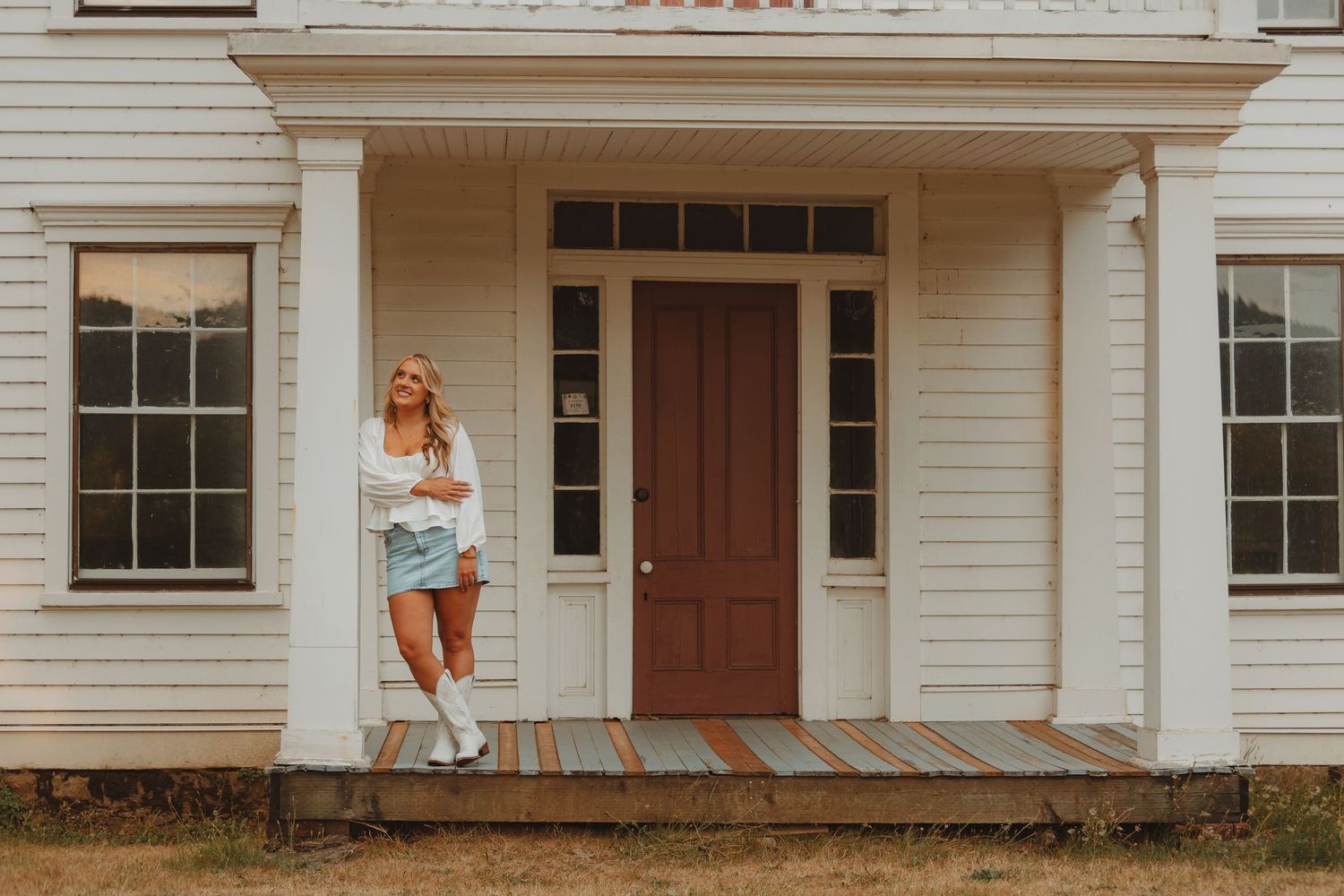 Woman in white top, denim shorts, and cowboy boots posing on porch of white farmhouse with red door.