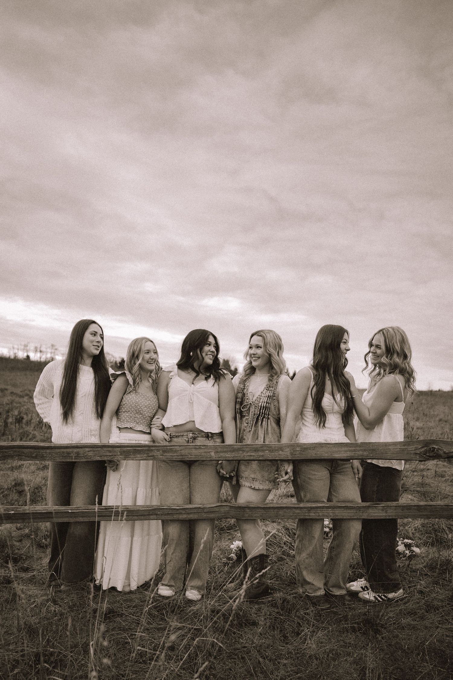 A group in white dresses and denim jackets poses along a wooden fence in a rustic outdoor setting.