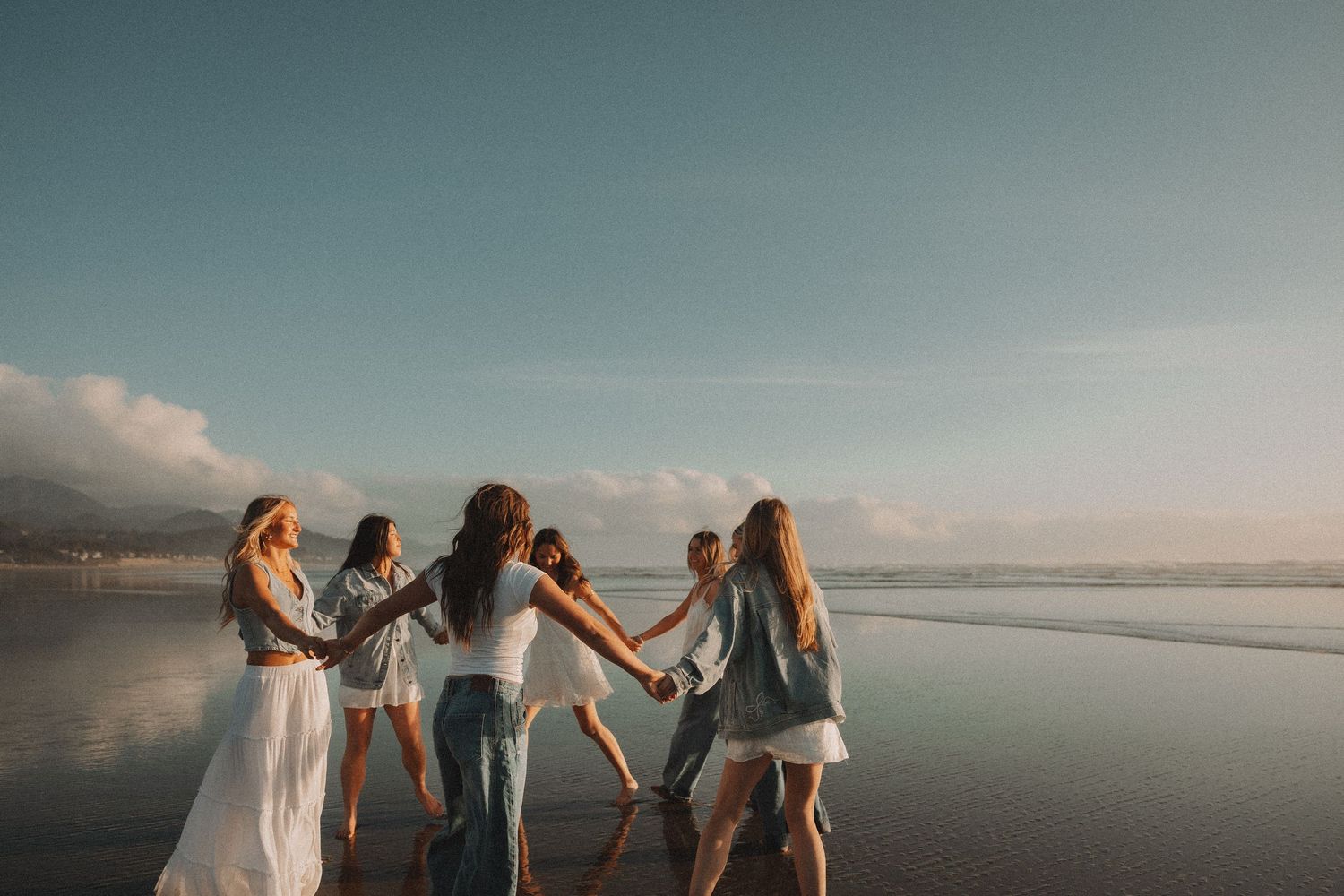 Six women holding hands in a circle on a beach at sunset, wearing casual outfits with ocean waves in the background.