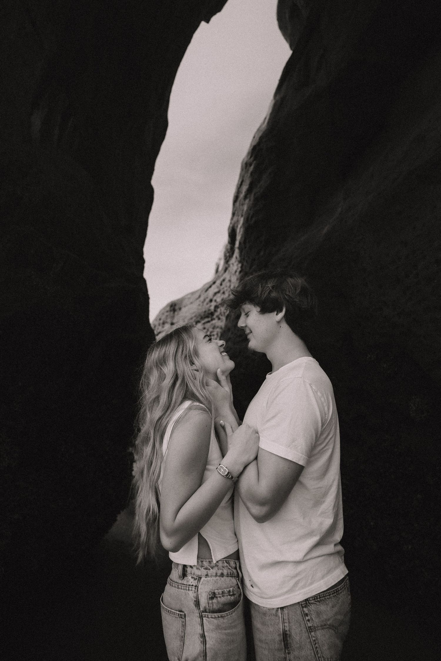 Black and white photo of a romantic couple standing close together in a rocky cave arch, about to kiss.