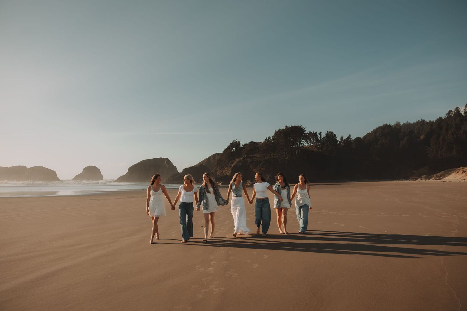 Eight women hold hands walking together across a wide sandy beach at golden hour.