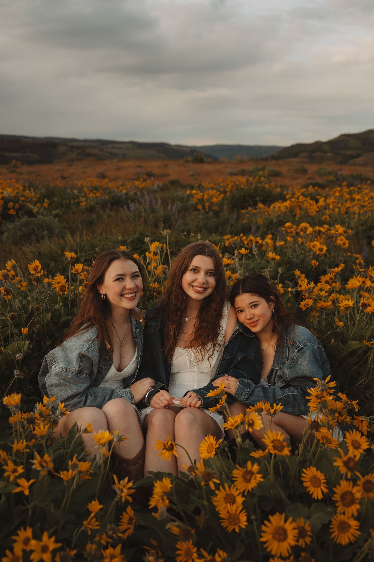 Three smiling young women sit together in a vibrant field of yellow sunflowers under a cloudy sky.