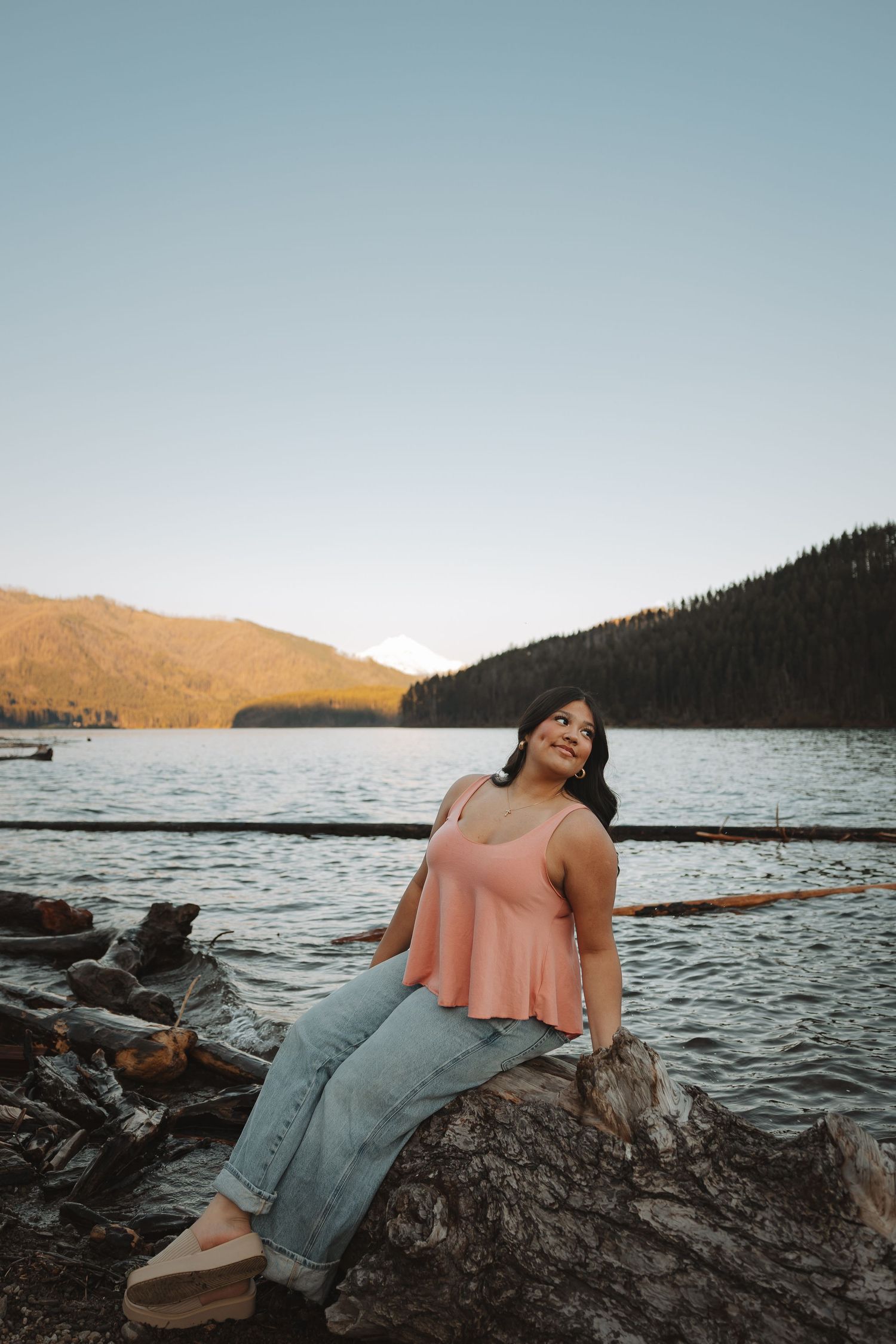 Young woman in pink top and jeans sitting on rocks by a serene mountain lake at sunset.
