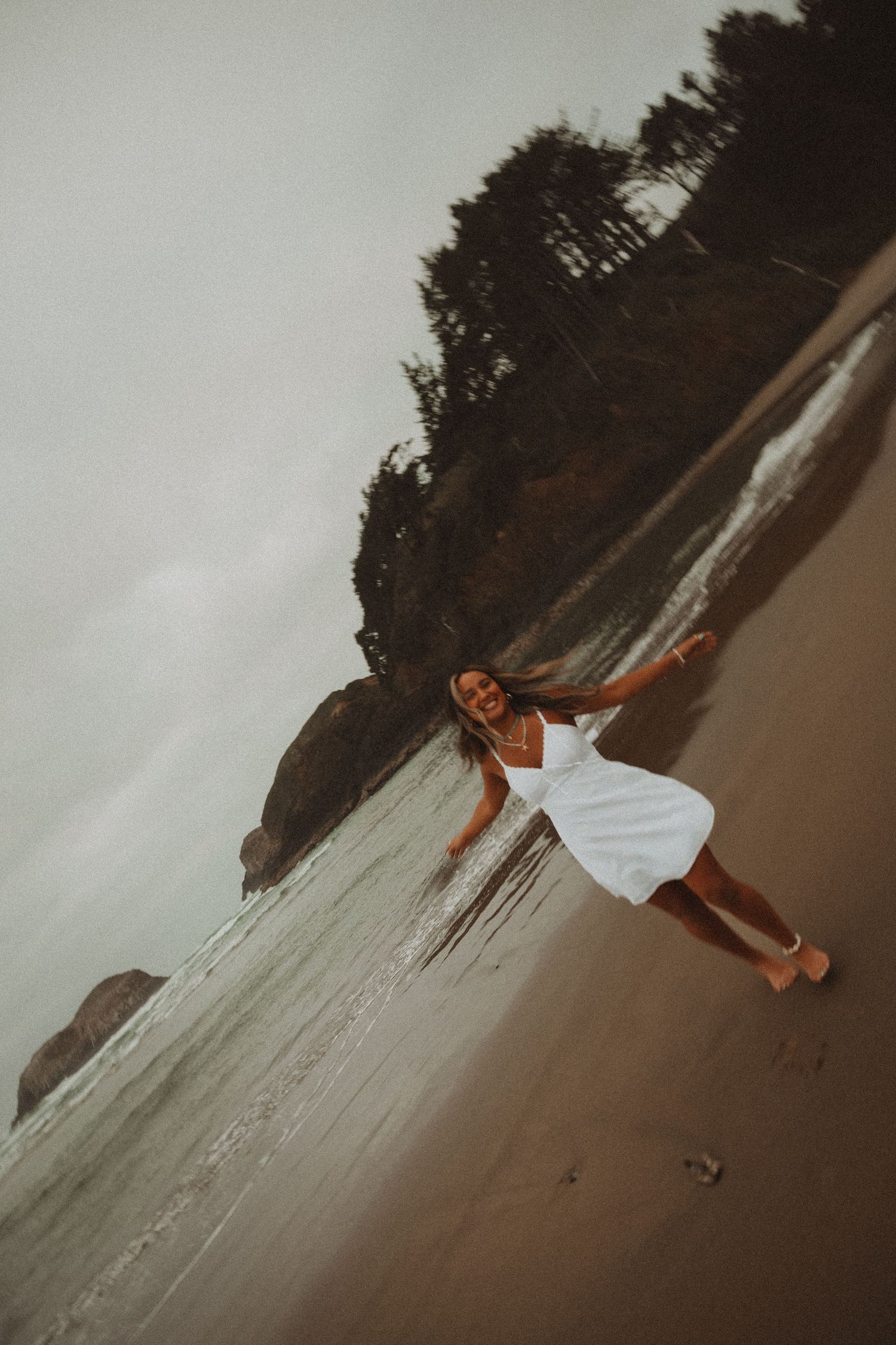 Woman in white dress laughing on sandy beach with ocean waves and rocky tree-lined shore in moody light.