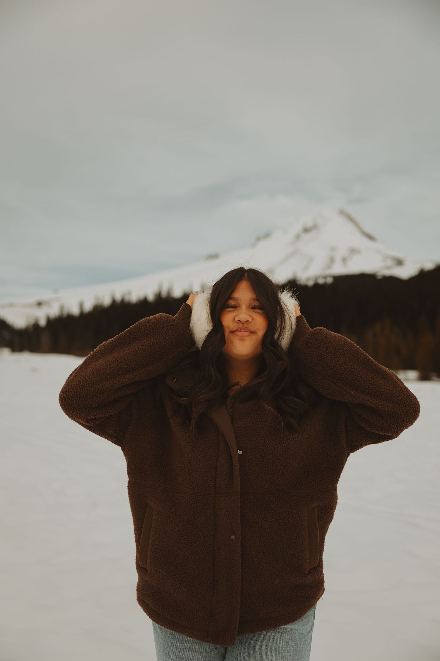 Young woman in brown jacket and white earmuffs smiling in snowy mountain landscape.