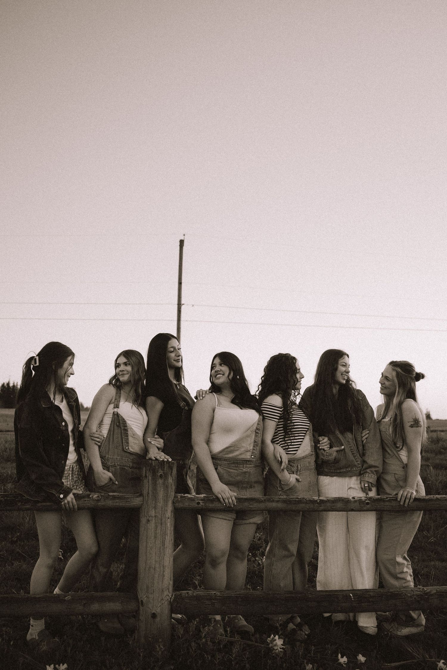 Seven young women laughing and leaning on a rustic wooden fence in a black and white photo.