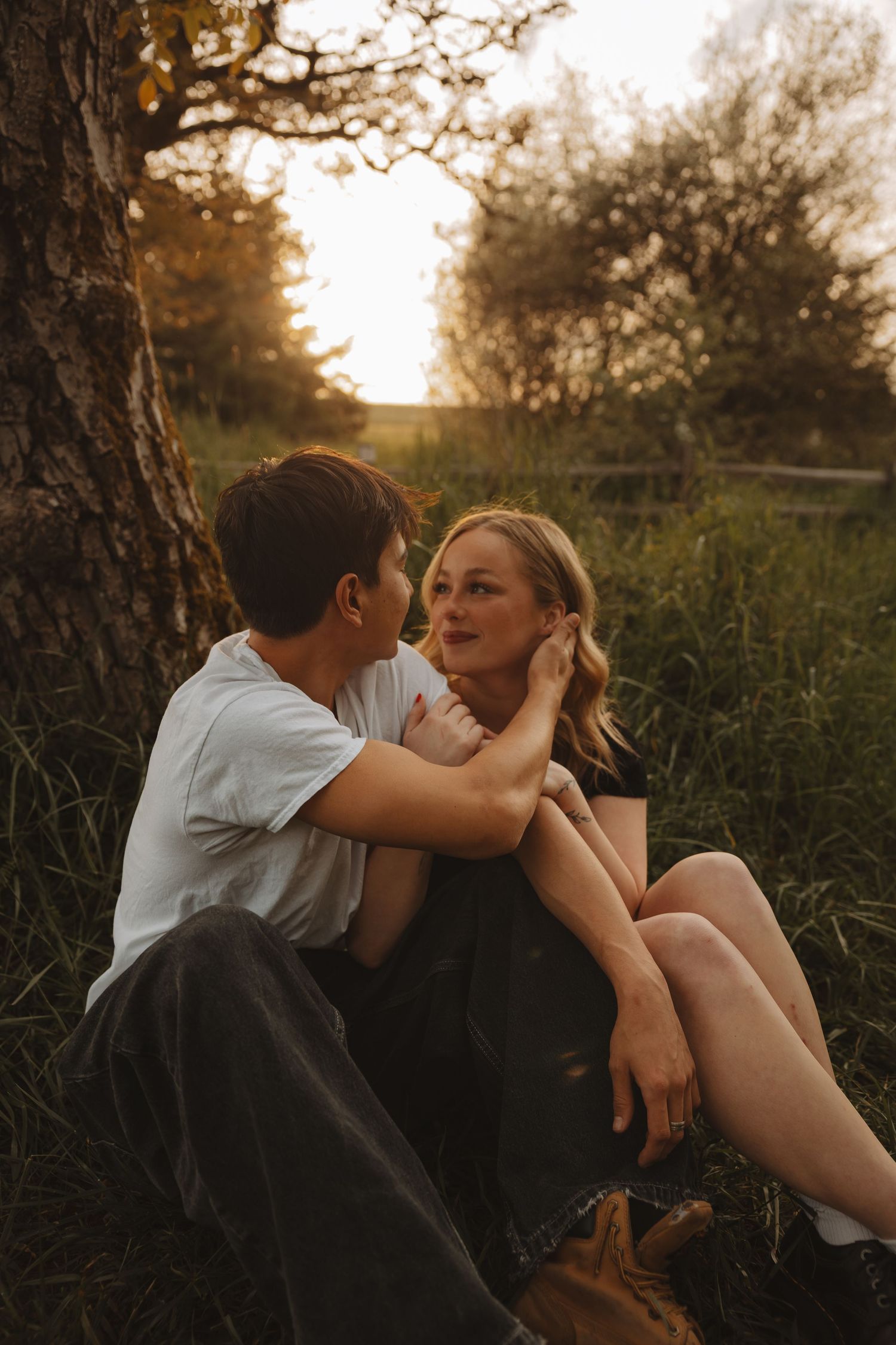 Couple sitting together at golden hour by a tree, man tenderly touching womans face in a romantic outdoor setting.