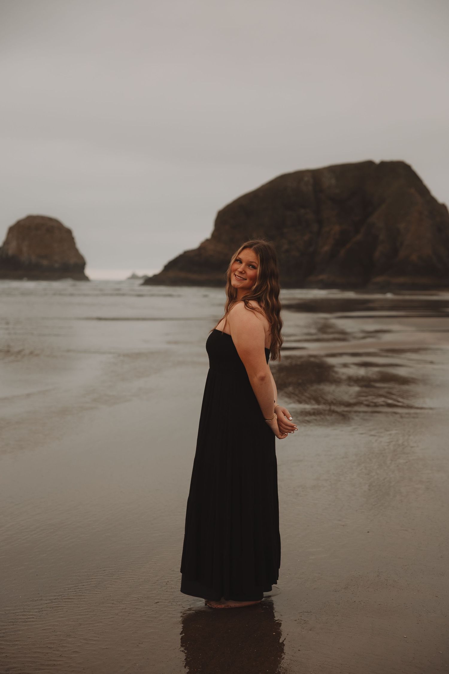 Person in black dress poses playfully on a moody beach with rocky cliffs in the background on an overcast day.