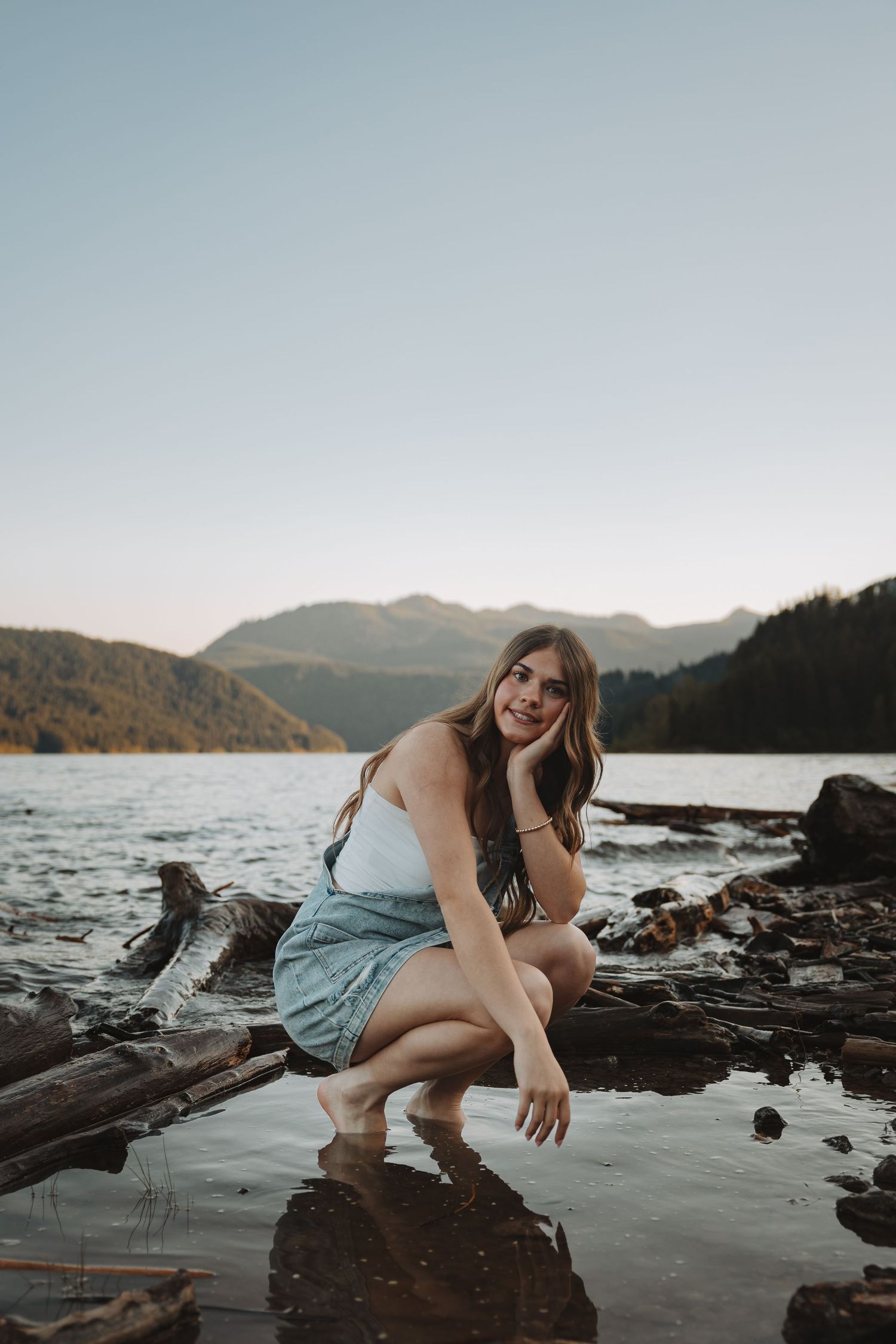 Young woman in white top and denim skirt crouching on driftwood by a mountain lake at sunset.
