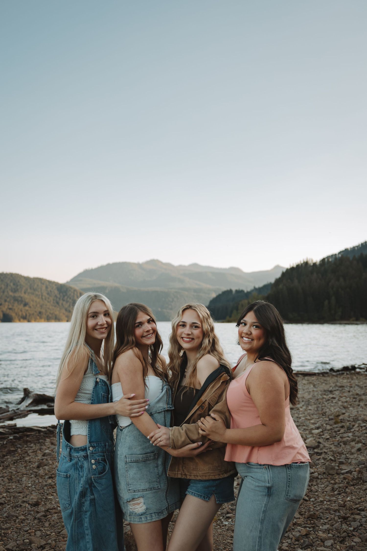 Four smiling young women posing together on a rocky lakeshore with forested mountains at sunset.